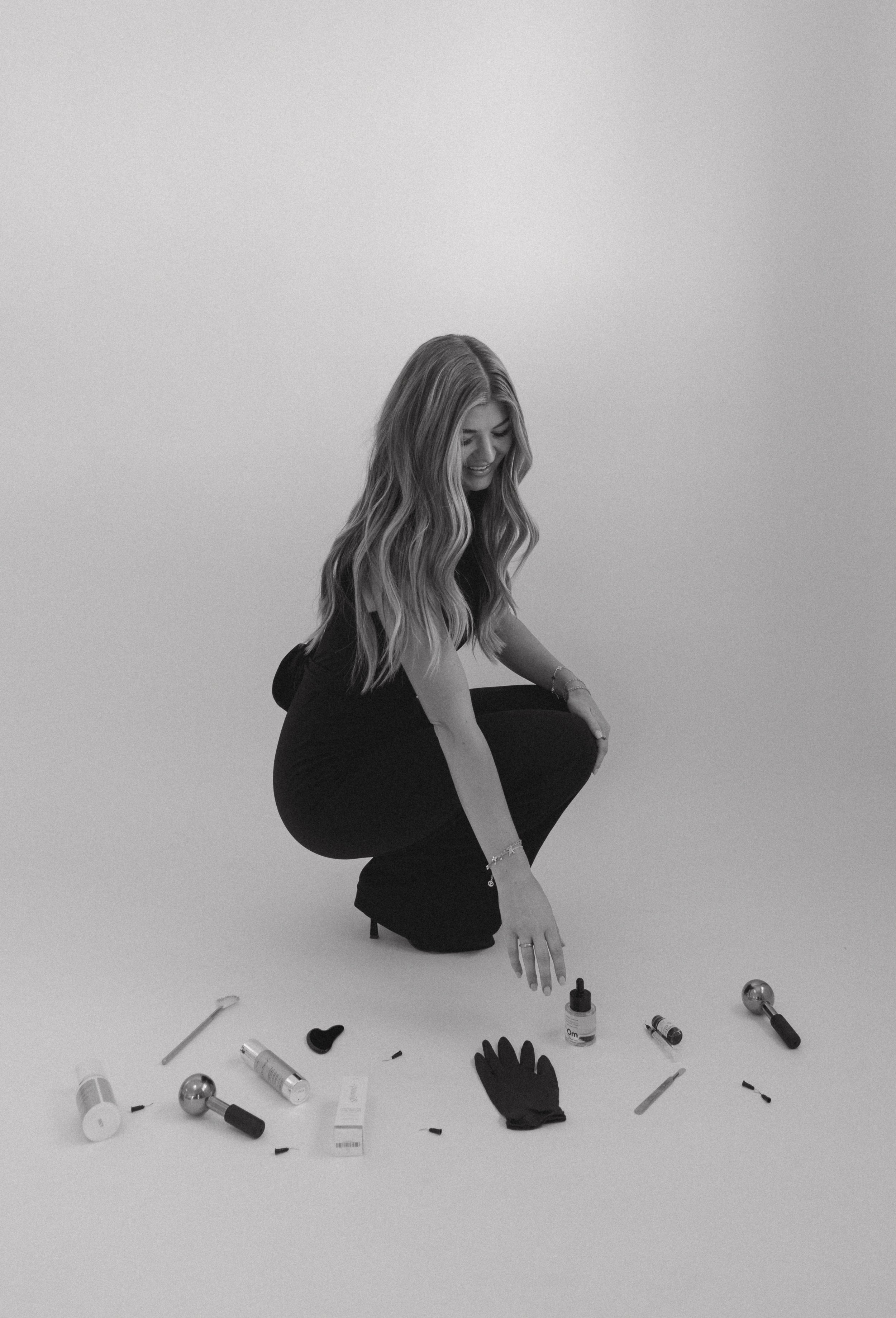 A woman with long wavy hair crouching on the floor, surrounded by various beauty or cosmetic products and tools, smiling while reaching for a small bottle.