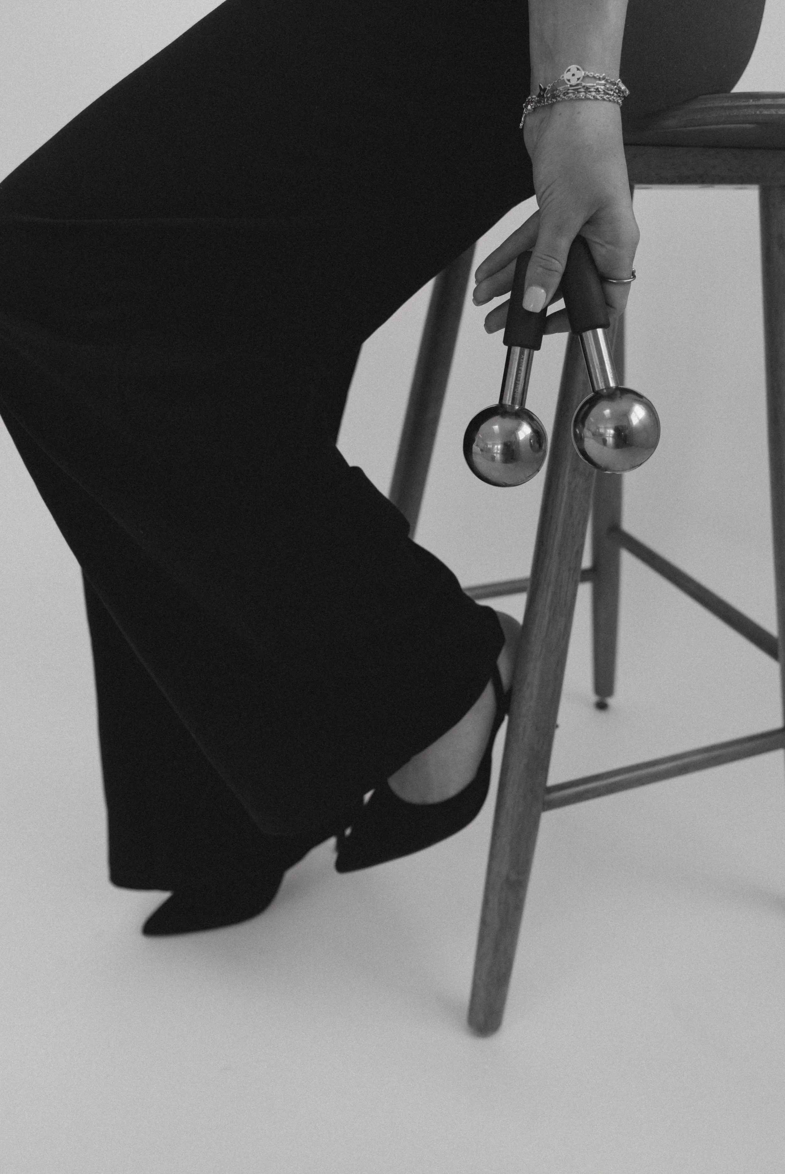 A person sitting on a wooden chair, holding two metallic massage tools, wearing black shoes with a cut-out design, and a bracelet, in a black and white photo.