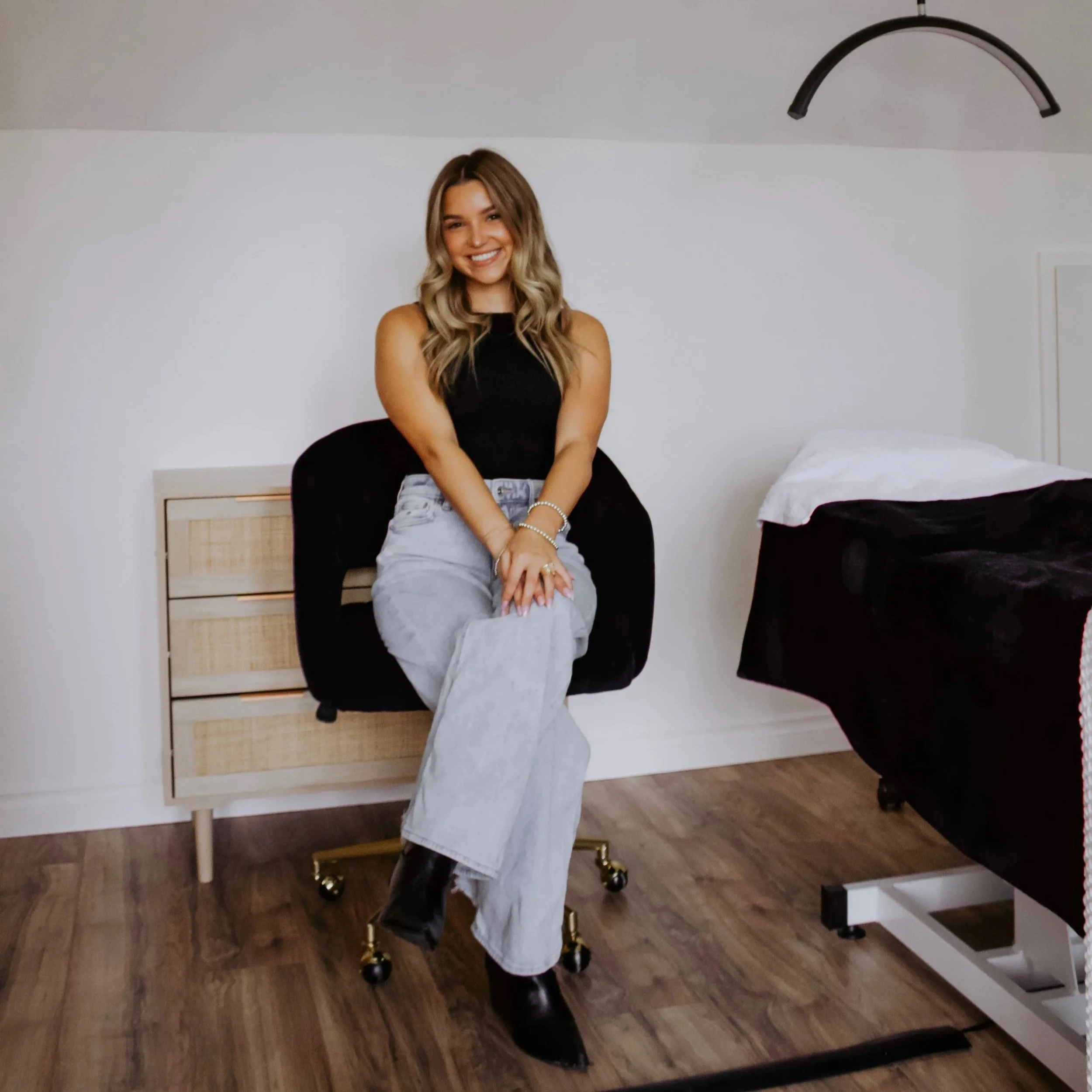 A young woman with long wavy hair, wearing a black sleeveless top, gray wide-leg pants, and black boots, sitting on a black chair with gold wheels in a room with white walls, wooden flooring, a beige nightstand with three drawers, and a bed with a black duvet and white pillow.