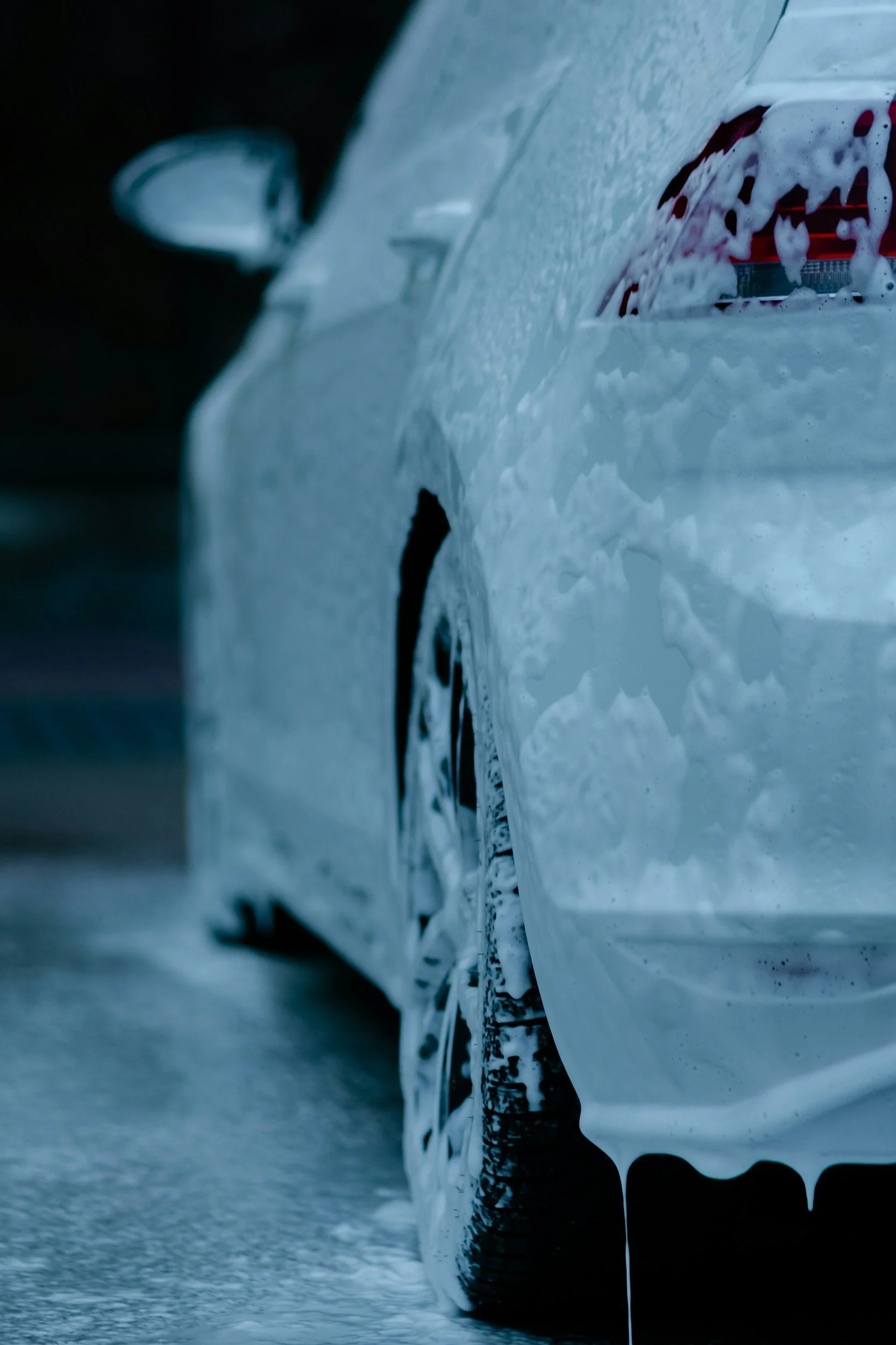 Close-up of a white car with soap and foam on its surface during a wash, focusing on the side panel and rear tire.