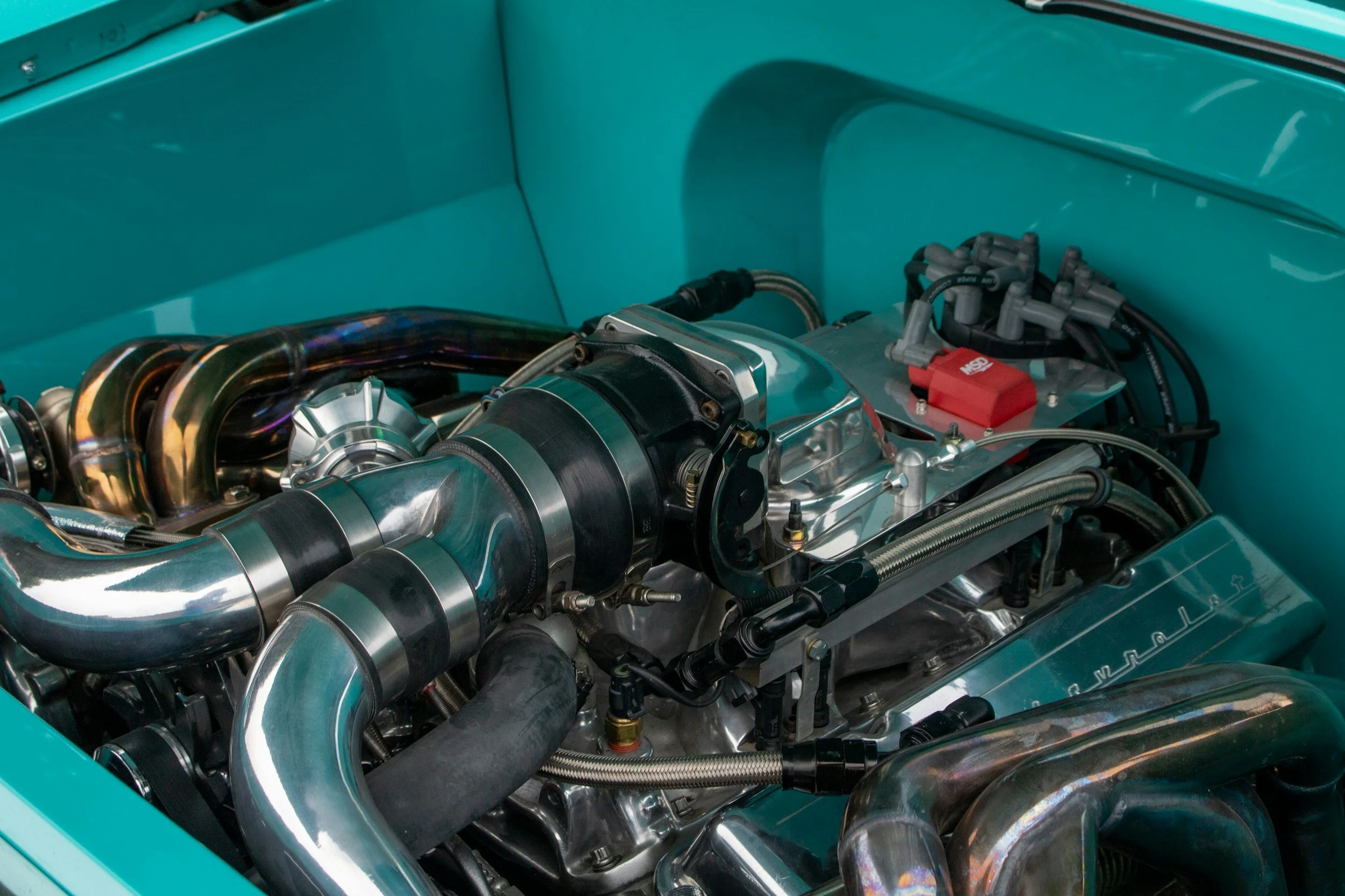 Close-up of a high-performance car engine with shiny metallic pipes and components in a teal-colored engine bay.