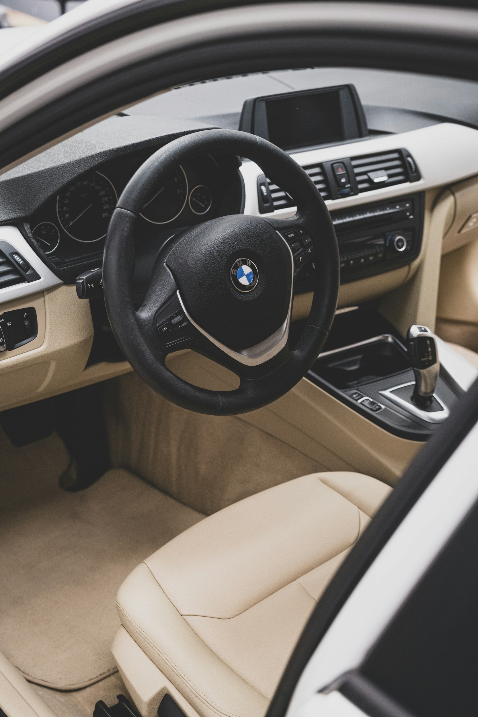 Interior of a BMW car showing steering wheel, dashboard, gear shift, and beige leather seat.