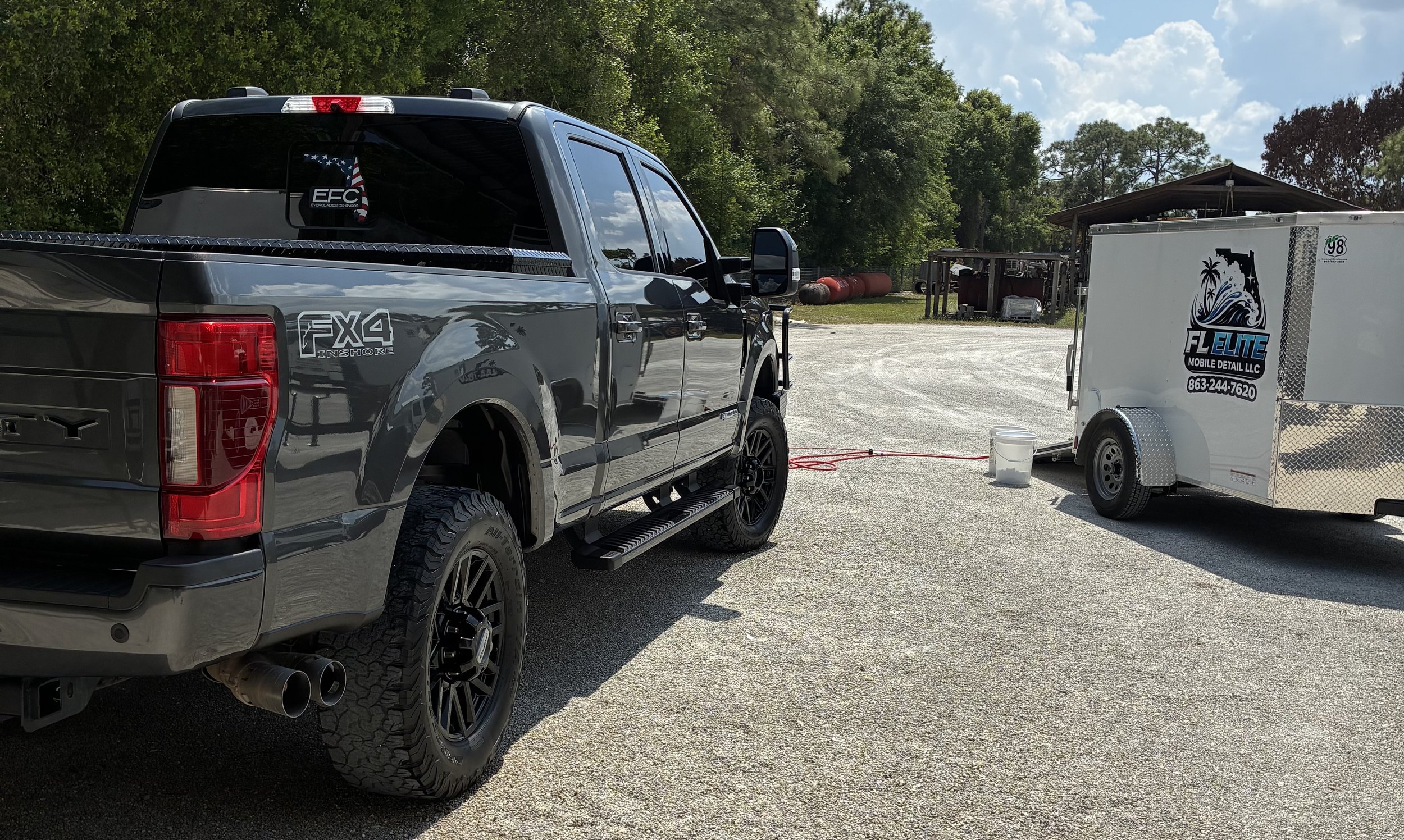 A black Ford pickup truck tied to a white mobile detailing trailer with a port and a bucket on the ground nearby, set in a gravel lot with trees and storage structures in the background.