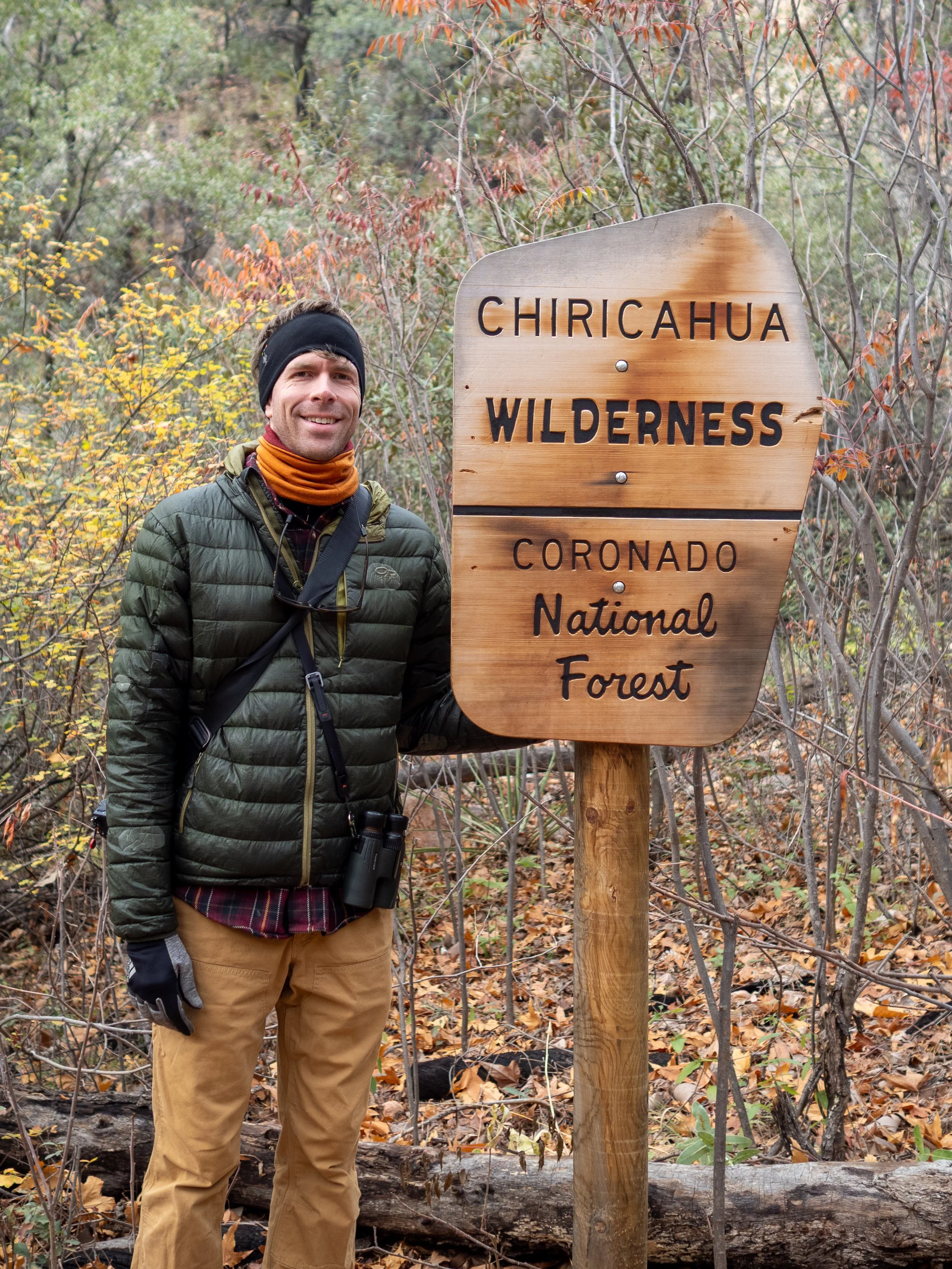 A man dressed in outdoor gear standing next to a wooden sign that reads 'Chiricahua Wilderness, Coronado National Forest', in a wooded area with fall foliage.