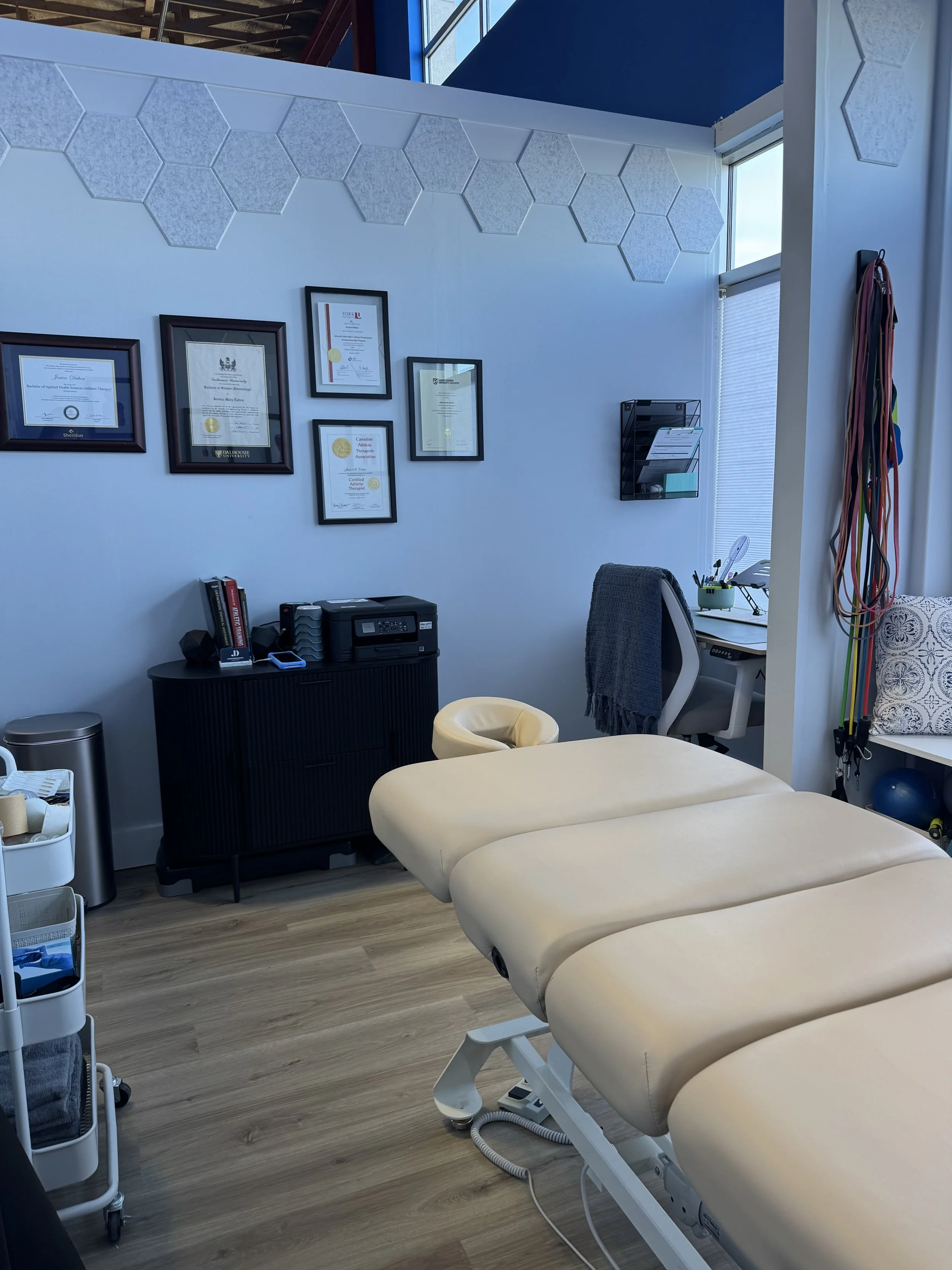 A therapy room with a massage table, framed certificates on the wall, a small black cabinet, a desk with a gray chair, a small round chair, and a wall-mounted organizer. There are exercise bands hanging on the side wall, and a window with blinds allowing natural light.