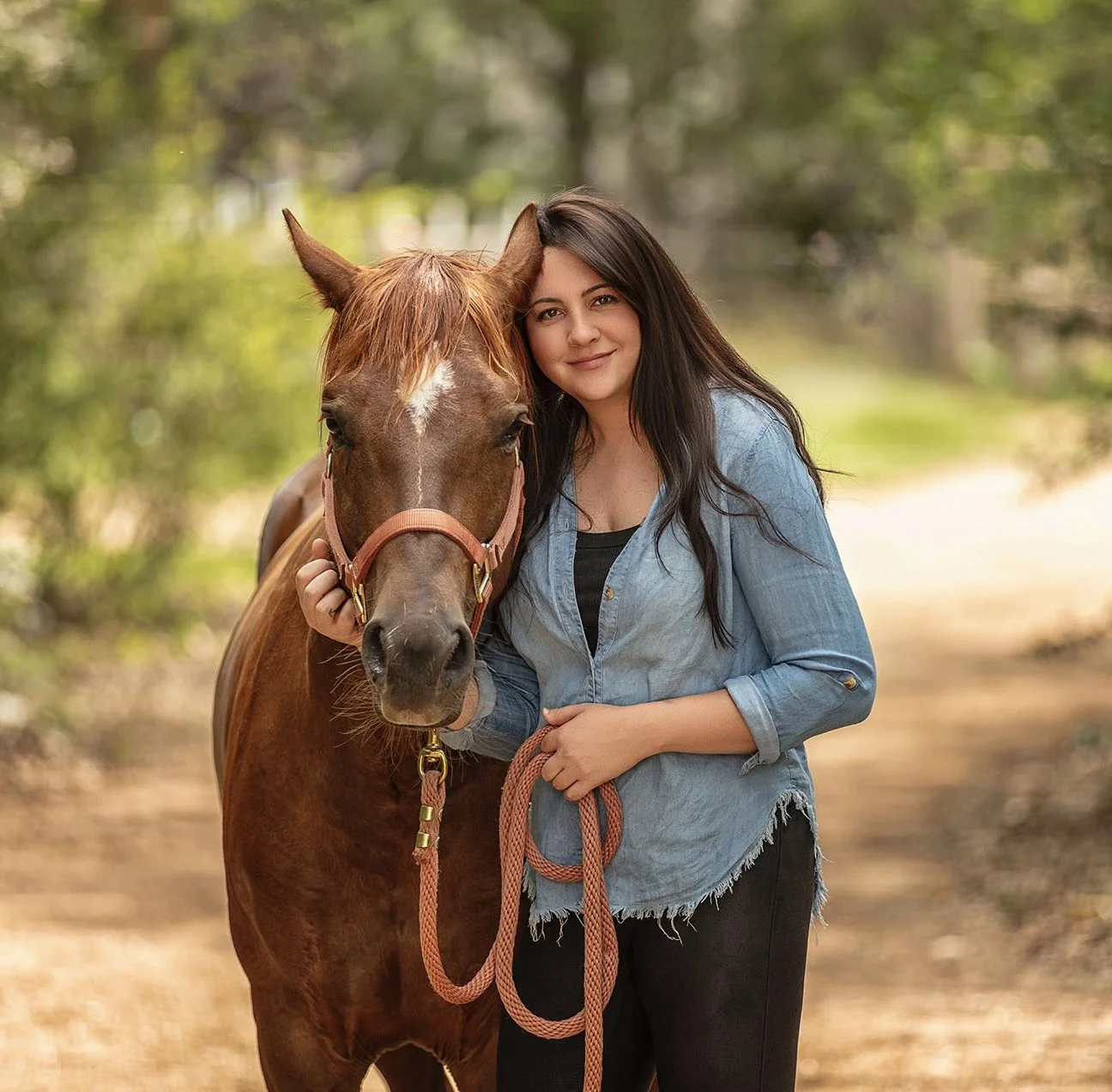 A young woman with long dark hair standing outdoors on a path in a wooded area, holding the lead of a brown horse, both looking at the camera.