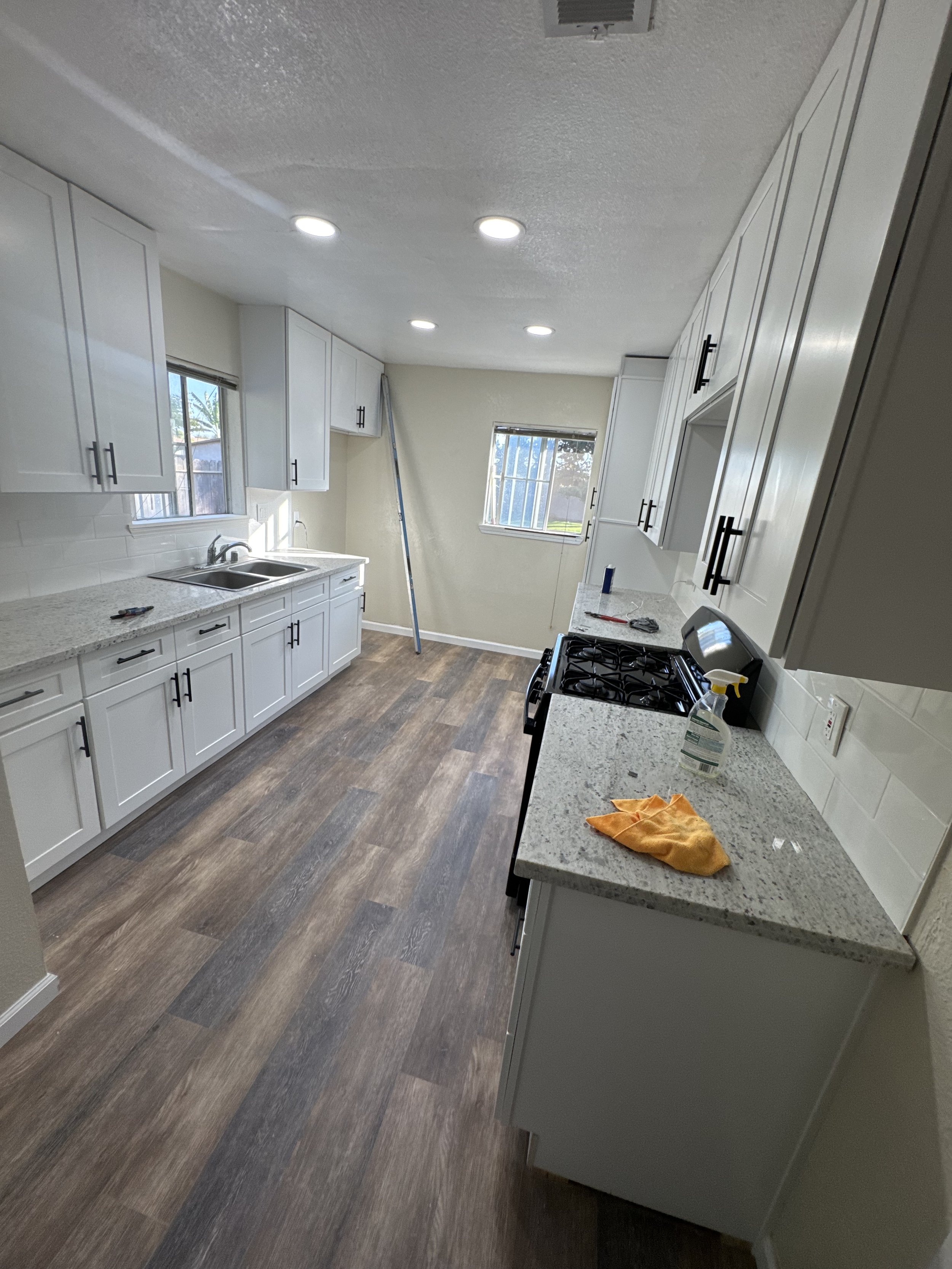 Kitchen with white cabinets, granite countertops, and dark wood flooring. There are two windows, a stove, and cleaning supplies on the counter.