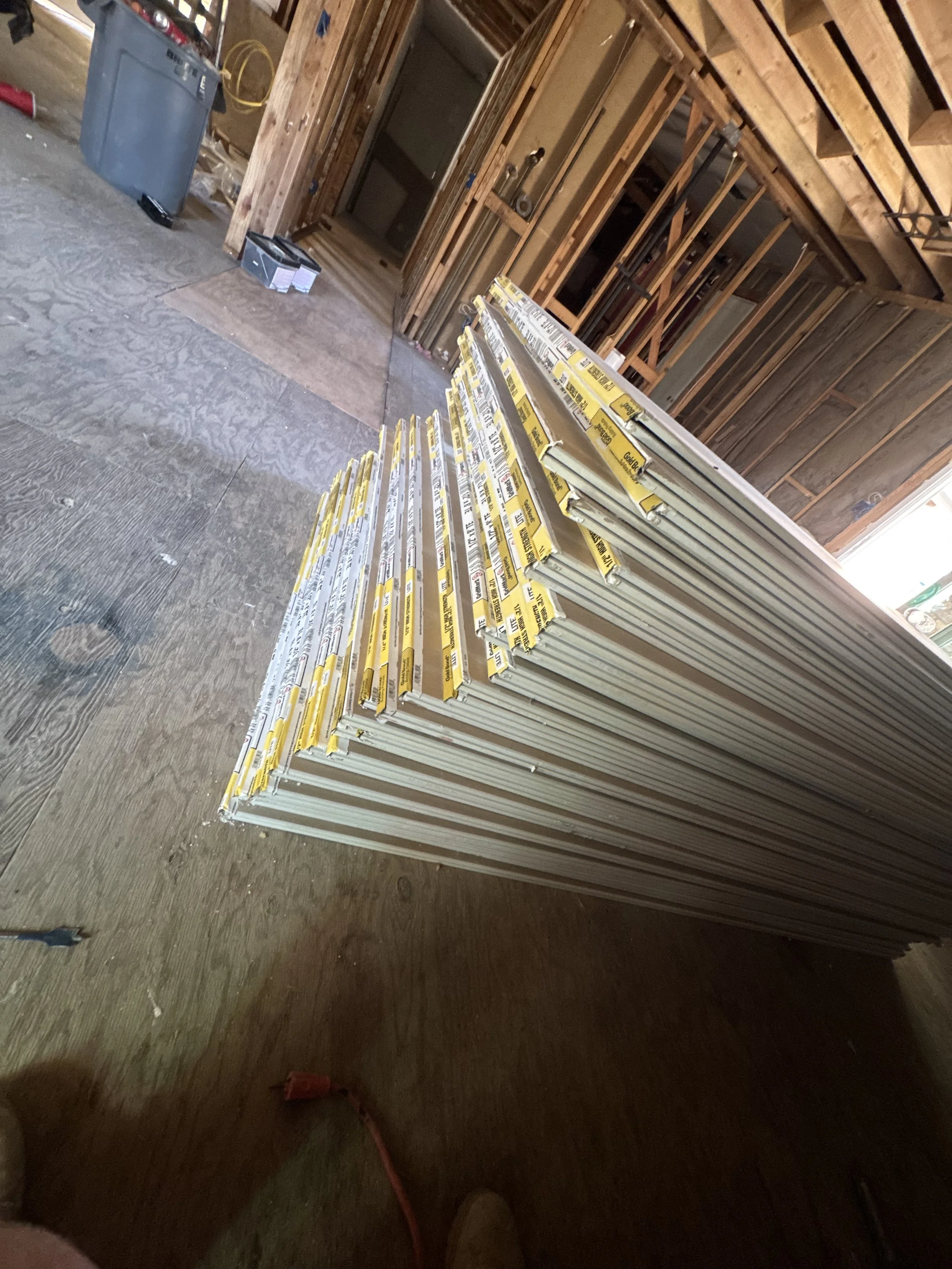 Stack of drywall panels in an unfinished room under construction, with wooden framing and construction tools visible.