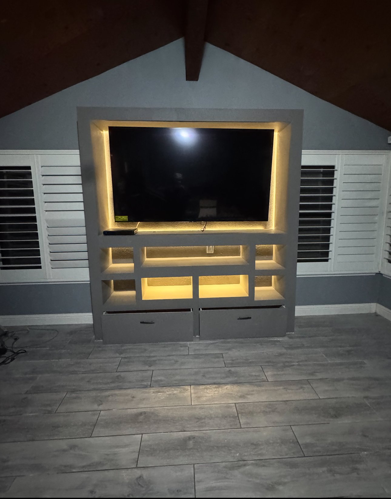 Living room TV unit with built-in lighting, a flat-screen TV, white and gray walls, and wood flooring.