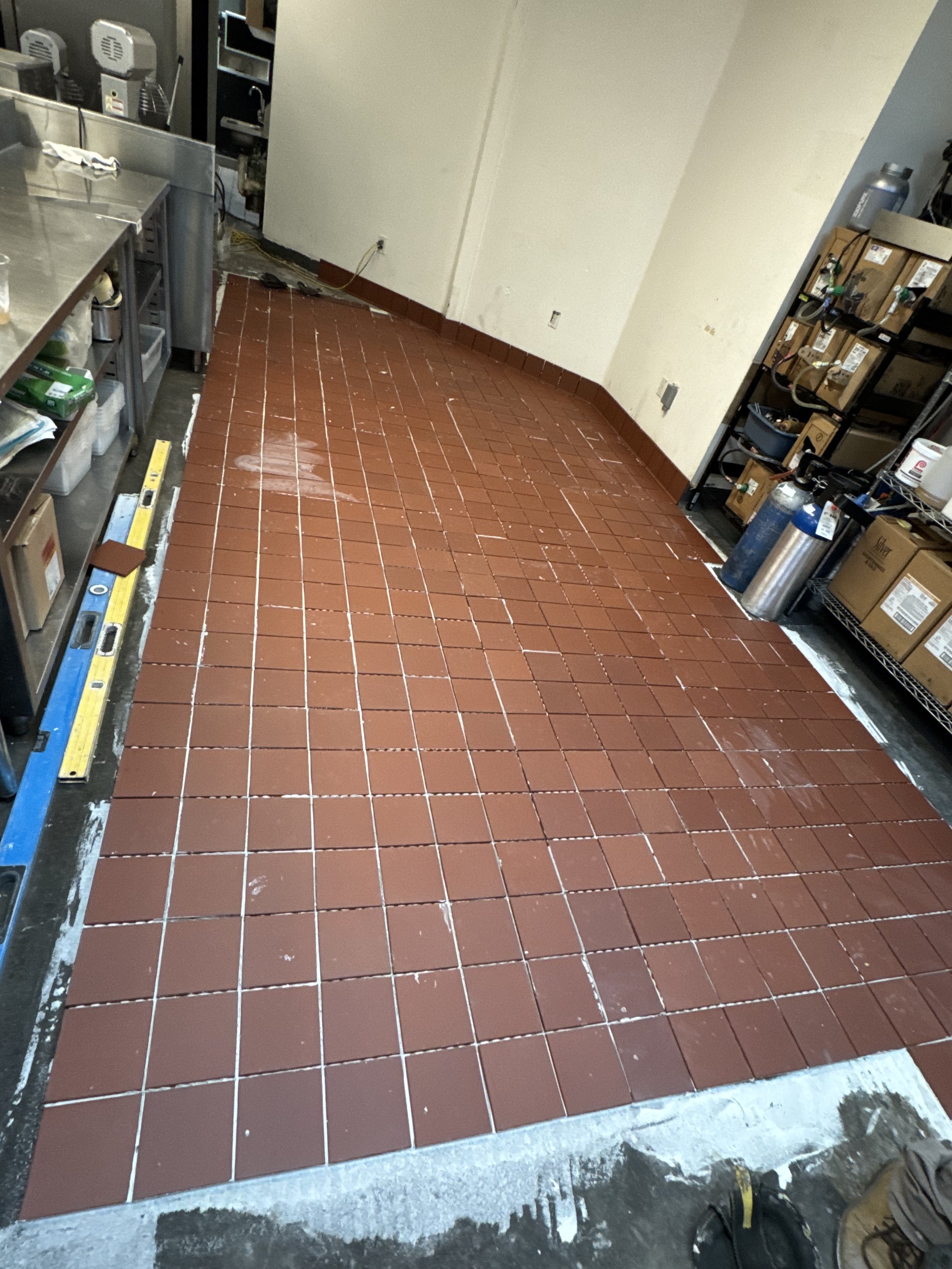 The floor in a kitchen is being tiled with new reddish-brown tiles; some tiles and grout lines are visible, and tools like a spirit level are on the floor. Shelves with boxes and equipment are on the right, and appliances are on the left.