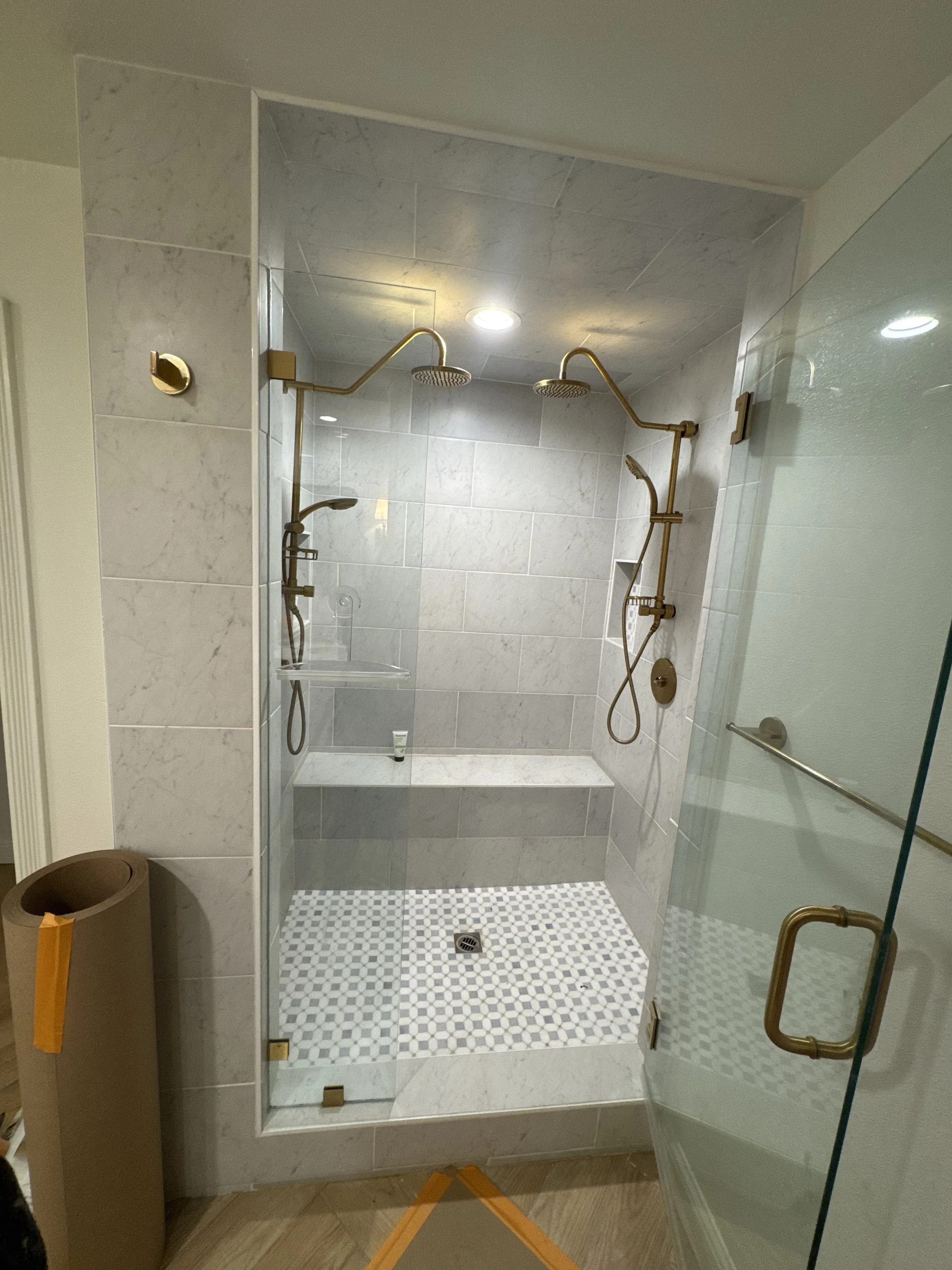 A walk-in shower with two rain shower heads and handheld shower attachments, tiled walls, a built-in shelf, and a glass door with gold hardware.