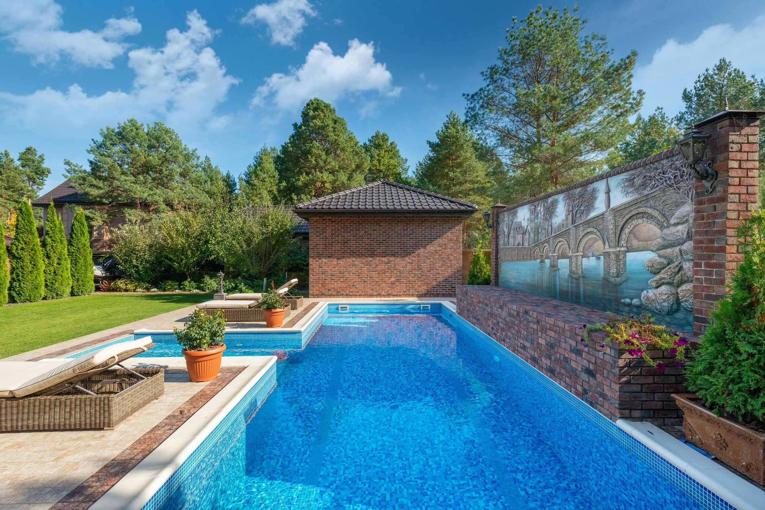 A backyard with a swimming pool, two lounge chairs, potted plants, a brick shed, a mural wall with a bridge scene, green grass, tall trees, and a partly cloudy blue sky.