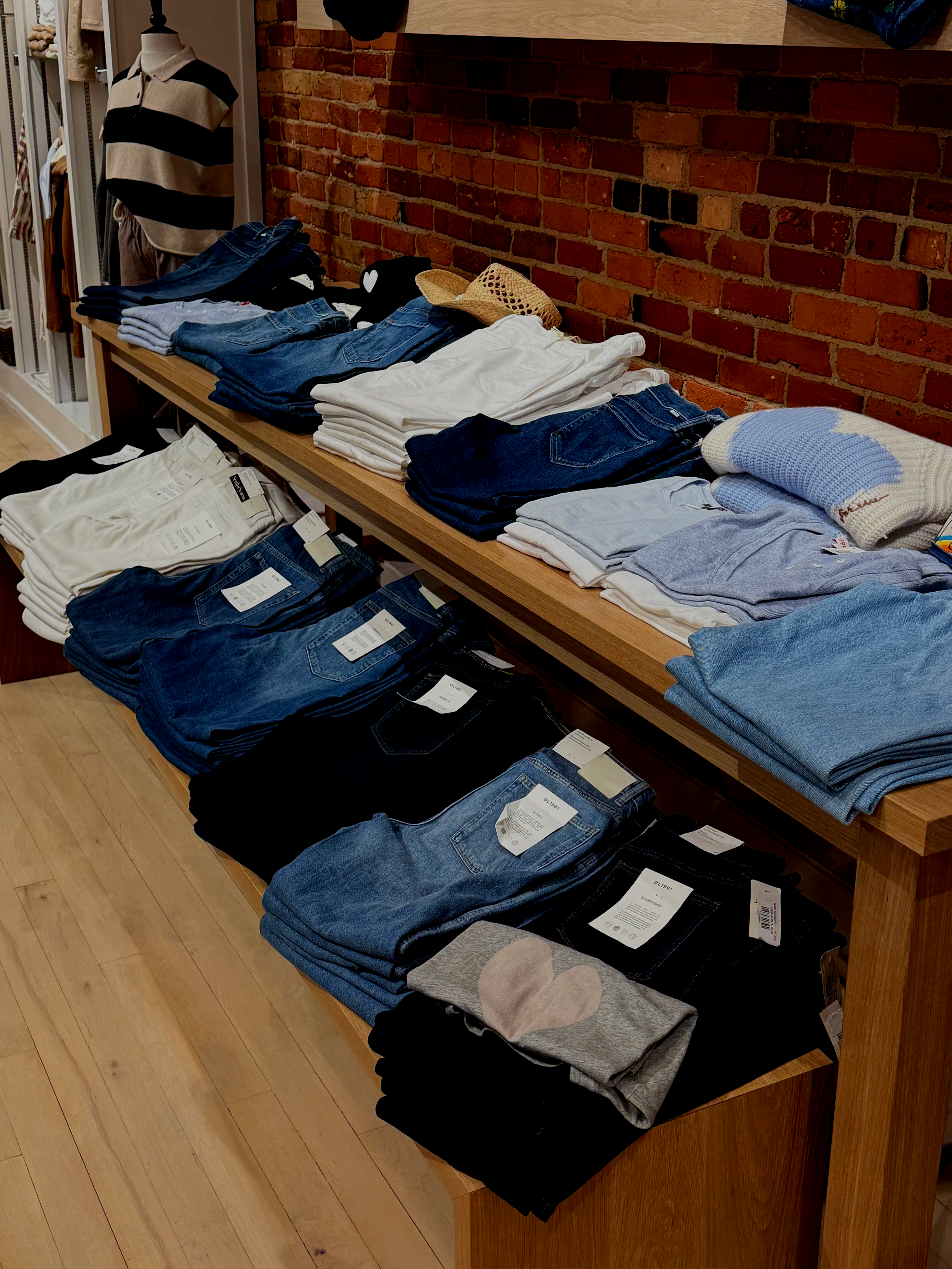 Display of men's clothing including folded jeans, T-shirts, and sweaters on a wooden table against a brick wall in a retail store.