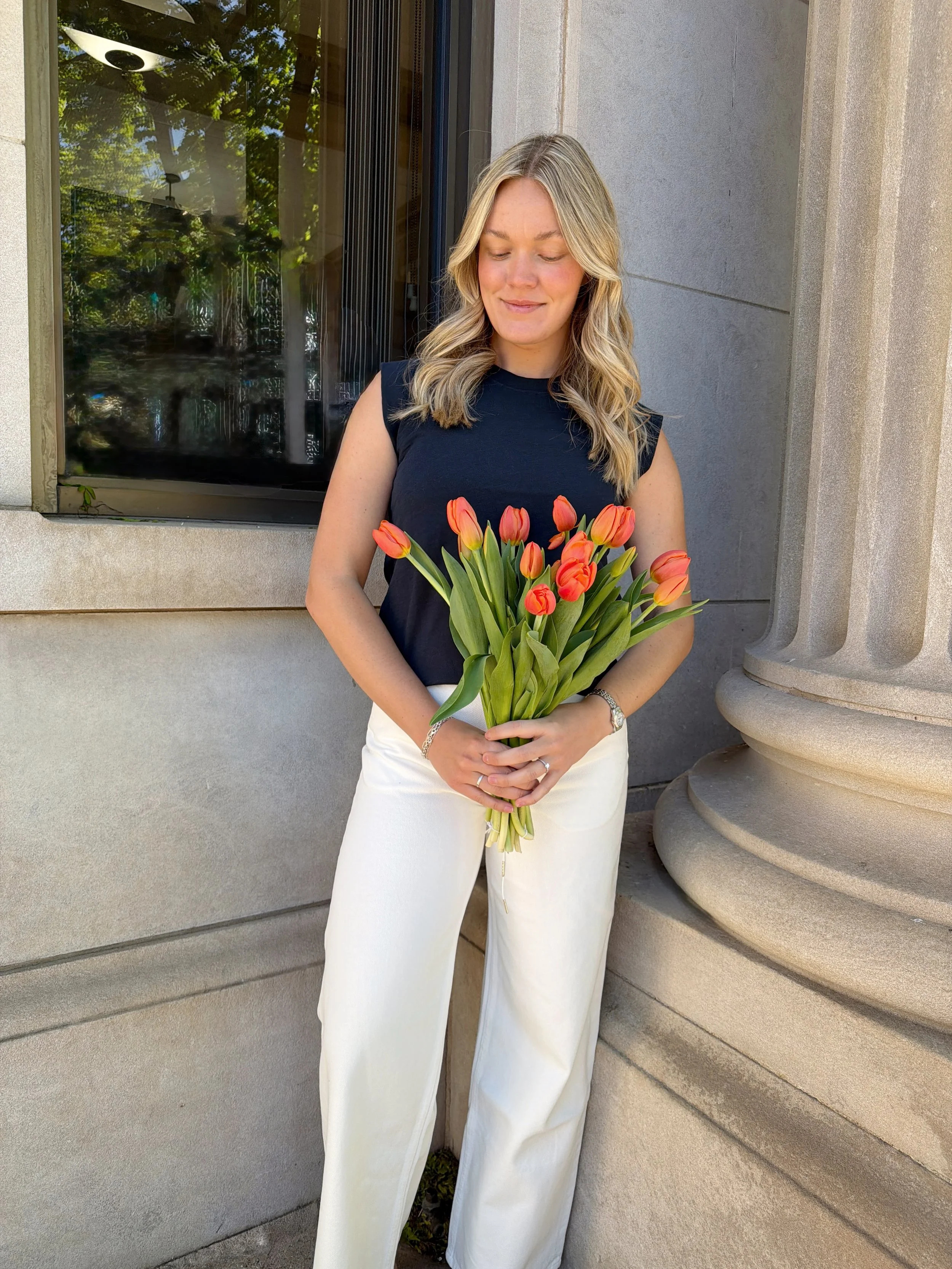 A woman with blonde hair in loose waves, wearing a black sleeveless top and white wide-leg pants, holding a bouquet of orange tulips, standing outdoors near a concrete building with a window and architectural columns.