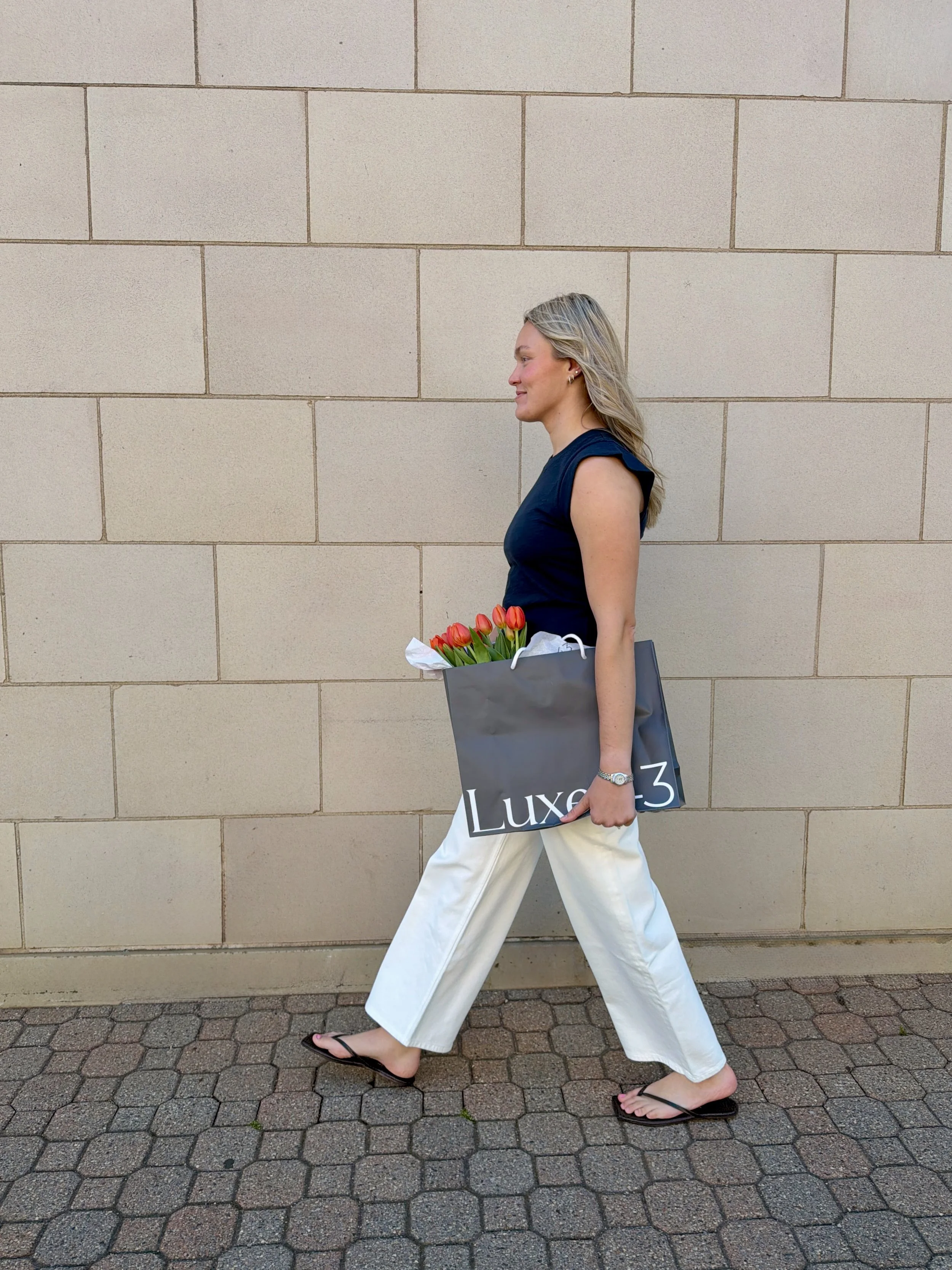 A woman walking on a cobblestone sidewalk in front of a beige brick wall, carrying a black shopping bag with red tulips inside.