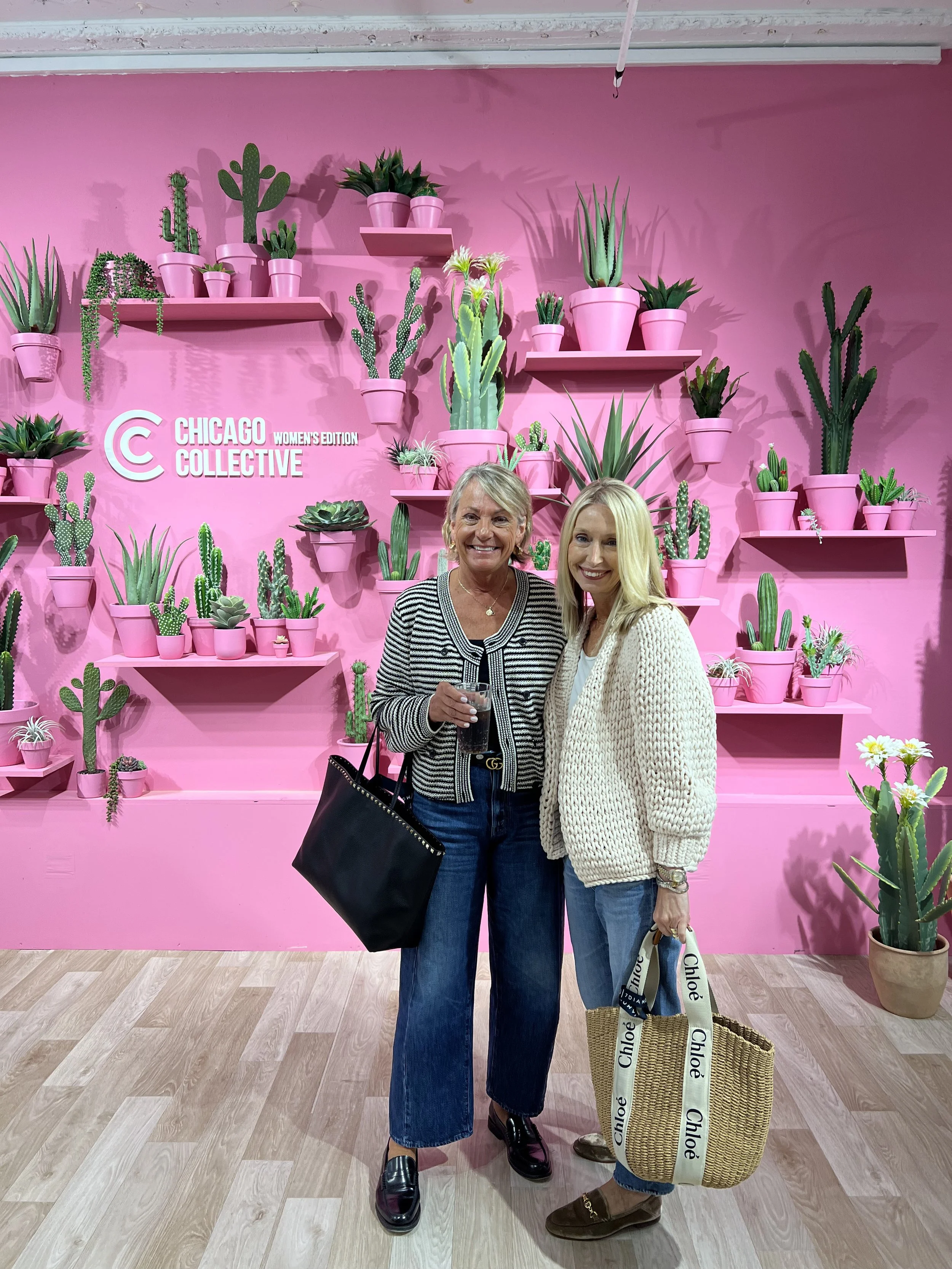 Two women standing in front of a pink wall with multiple potted cacti and succulents, and a sign that says 'Chicago Women's Edition Collective.' They are smiling and holding drinks and a bag, dressed in casual outfits.