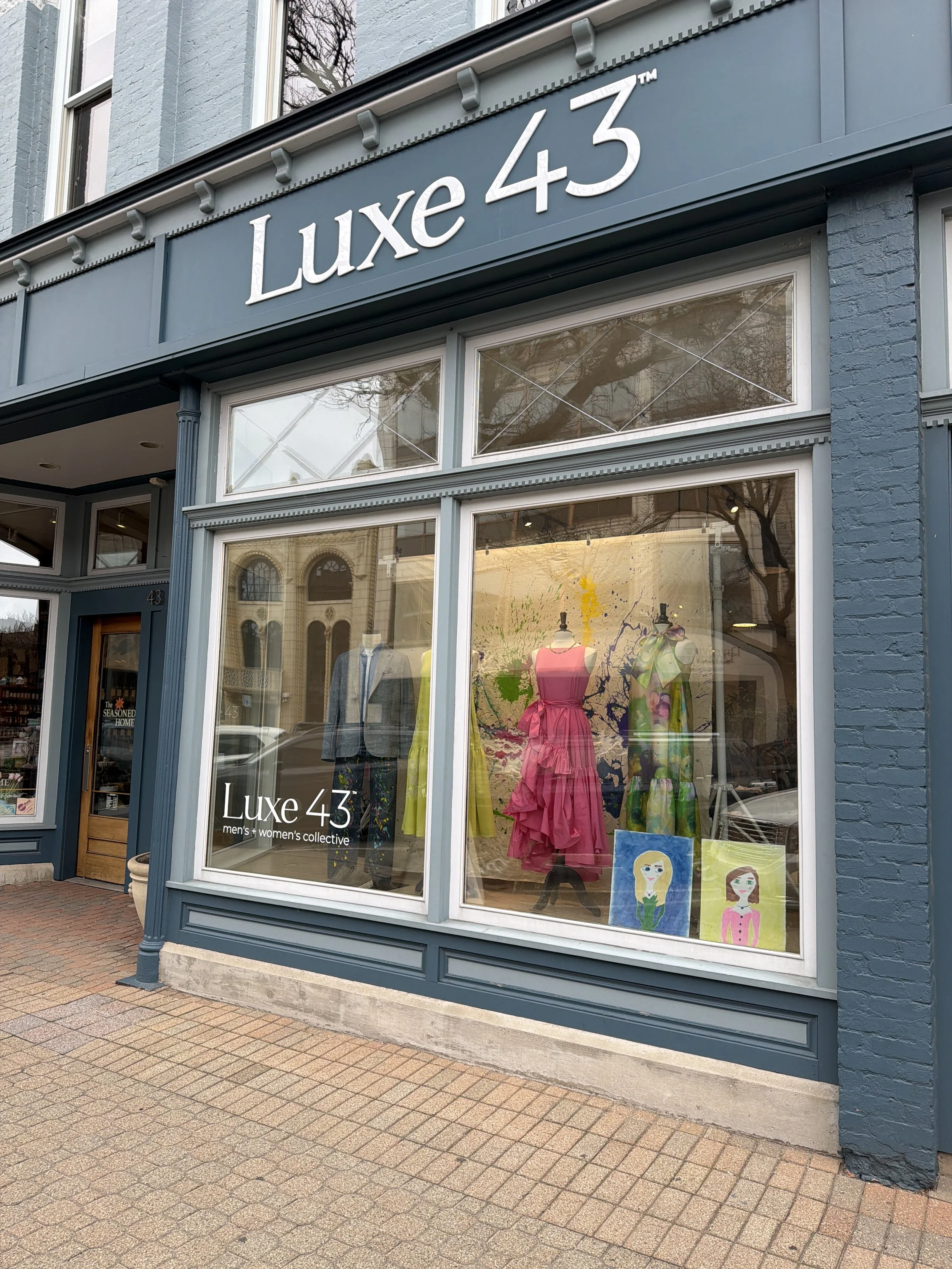 The storefront of Luxe 43, a men's and women's clothing store, showing mannequins dressed in colorful dresses and artwork displayed in the large display window.