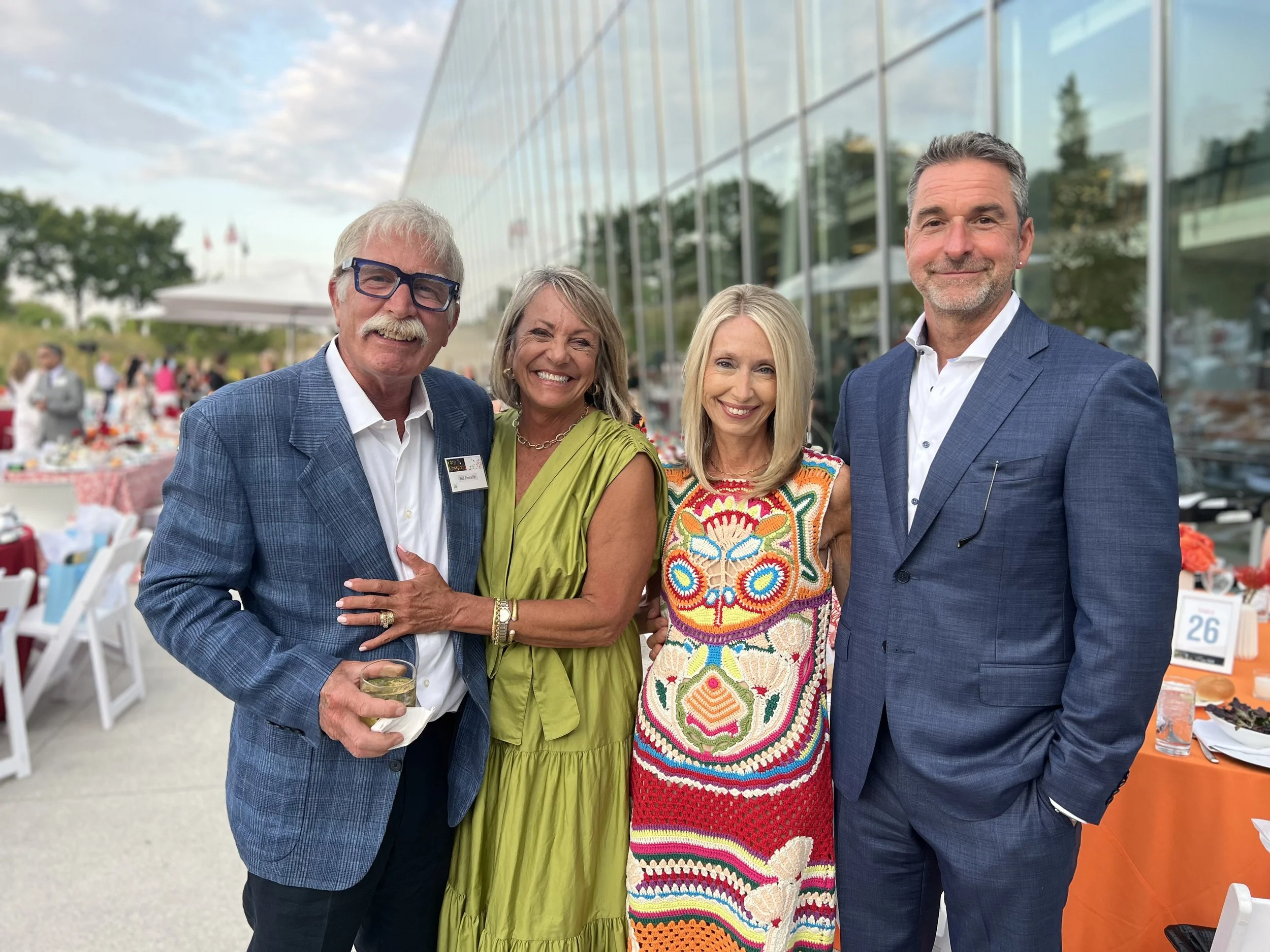 Four people, two men and two women, standing together outdoors in front of a modern glass building, smiling, dressed in business or semi-formal attire at a social event with tables and chairs in the background.