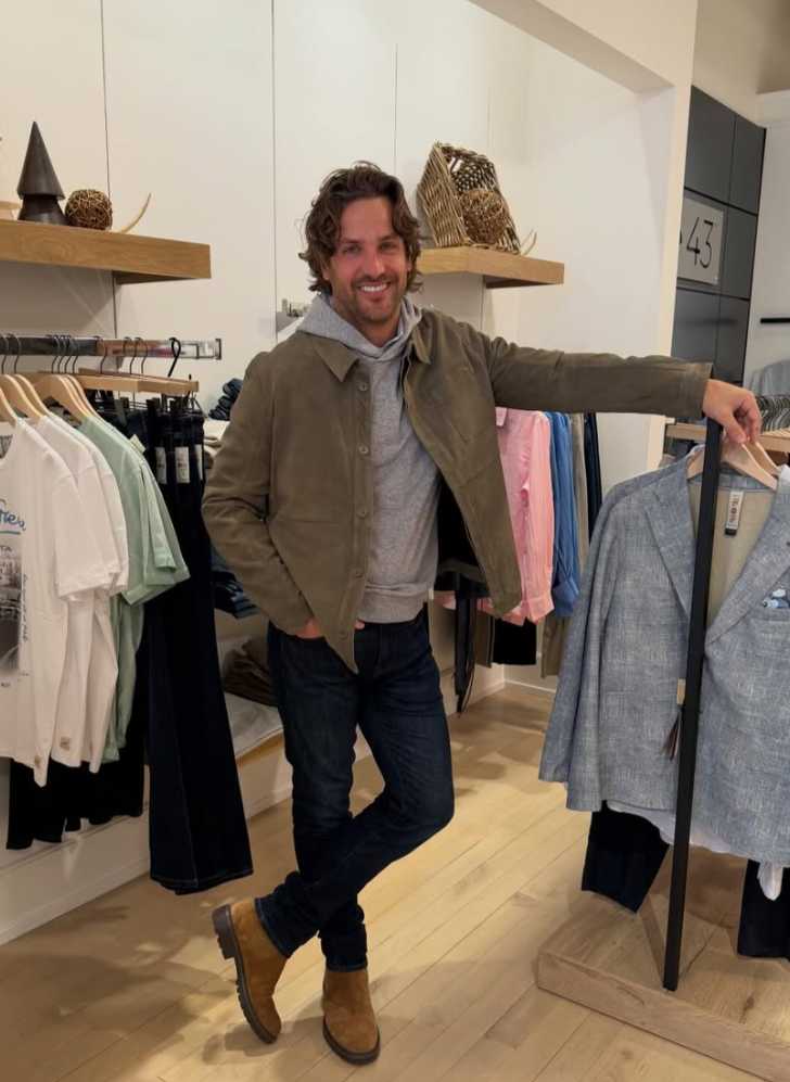 A smiling man with curly hair, wearing a light gray hoodie, a brown jacket, and dark jeans, standing in a clothing store, holding a gray blazer on a hanger.