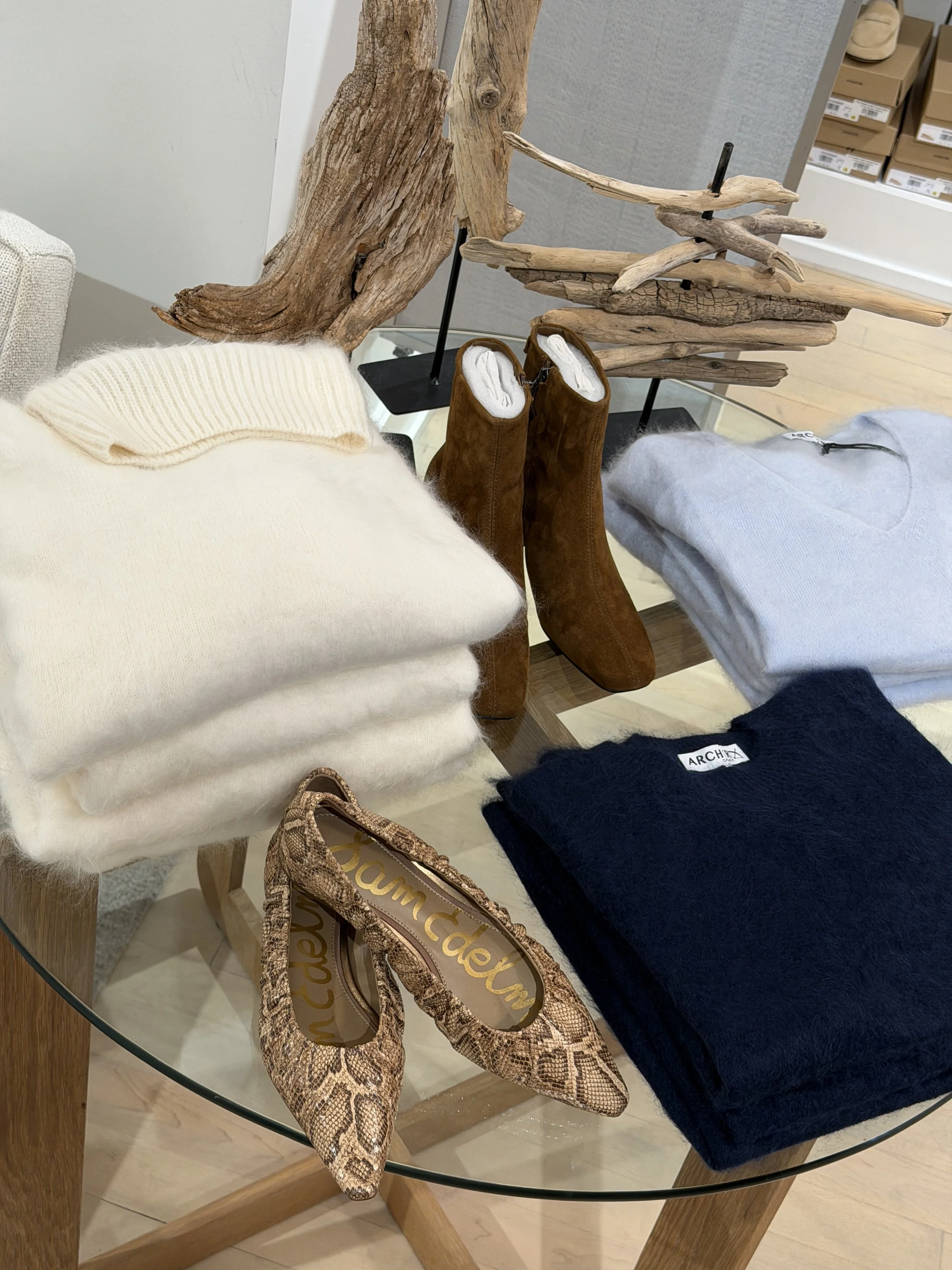 A table with an archer cream sweater and two archer blue and navy sweaters with a pair of brown suede sam edelman boots and a pair of pointed toe snake skin sam edelman flats.