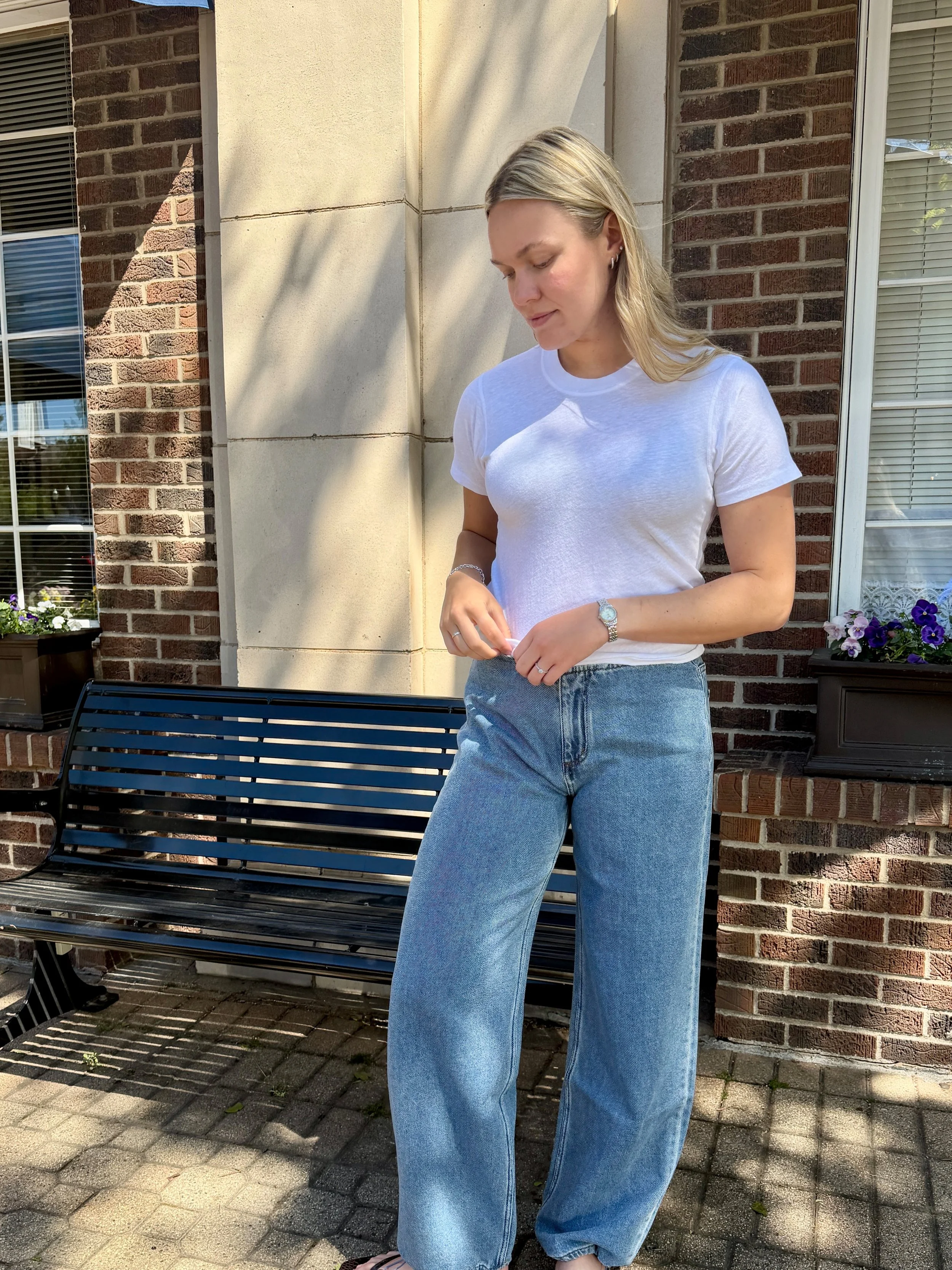 A woman with blonde hair wearing a white T-shirt and blue jeans standing outside in front of a brick building with windows, a black bench, and a window box with purple, white, and yellow flowers.