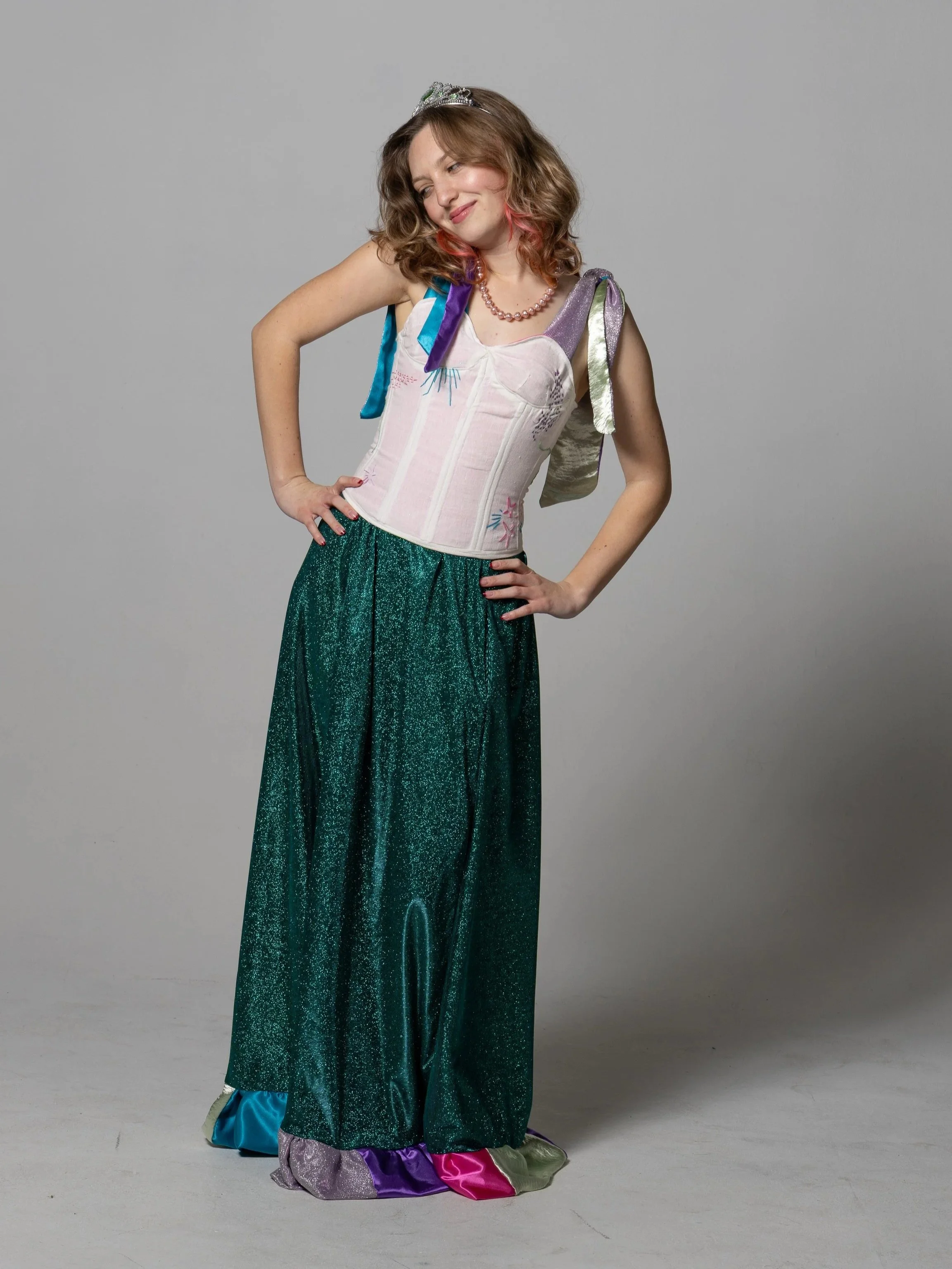 Daylin wearing a tiara, a pearl necklace, a colorful sleeveless top, and shiny, multicolored, glittering skirt, standing with her hands on her hips and smiling.