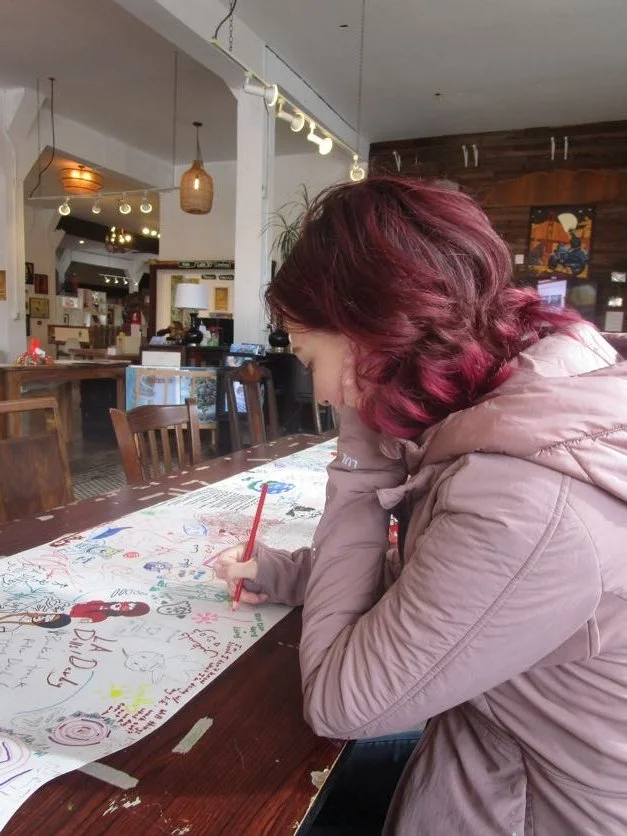 A woman with purple hair drawing on a large piece of paper with a red marker, sitting at a wooden table in a cozy café eclectic decor.