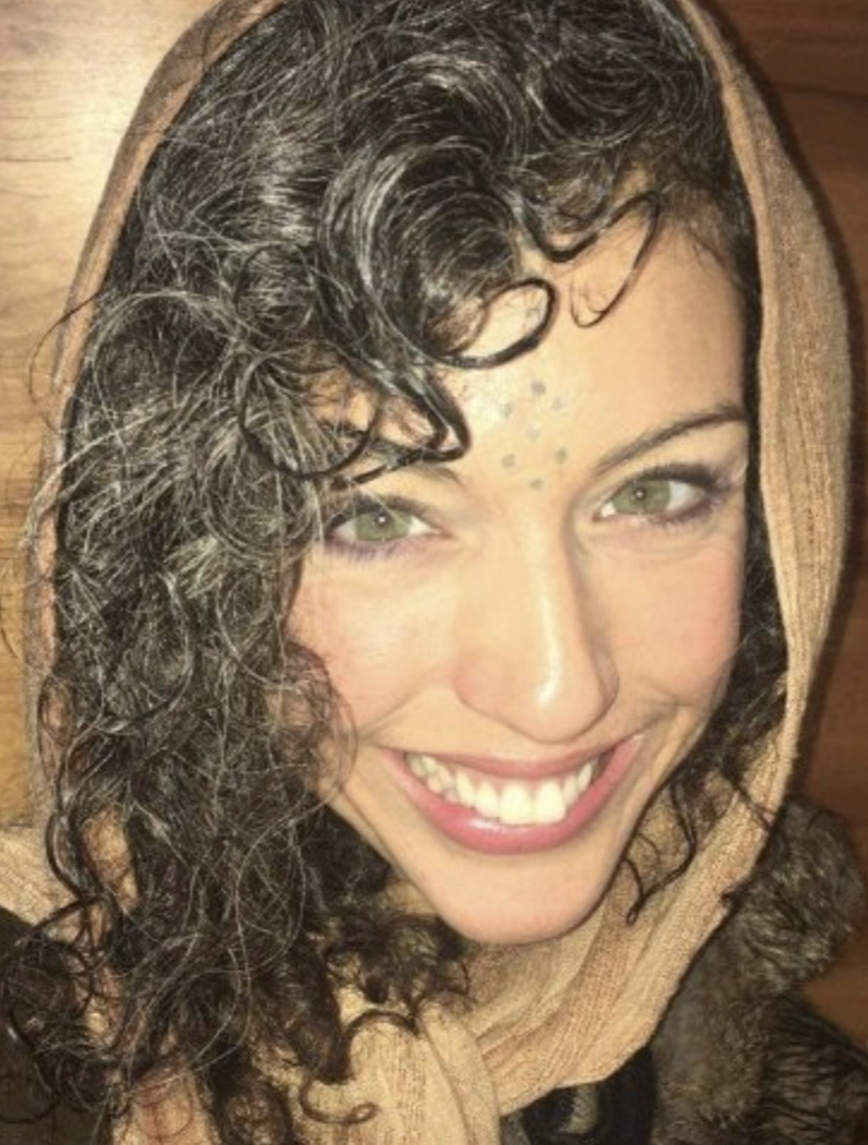 A woman with long, wavy, blonde hair smiling while holding a pair of black headphones in a cozy, artfully decorated room with wooden walls, a skeleton model, bookshelves, and a bed with a yellow blanket.