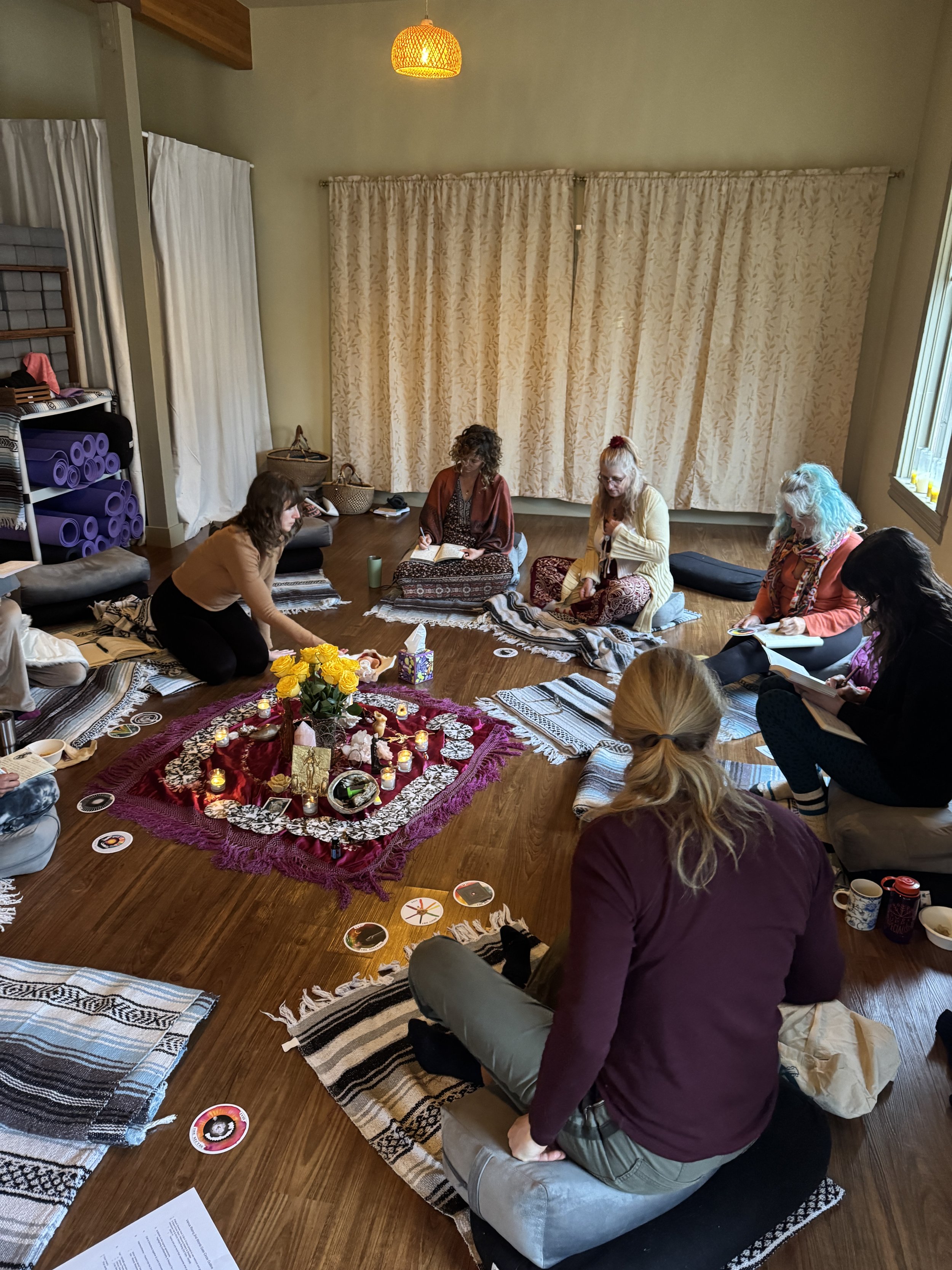 Group of women sitting on the floor in a circle, participating in a spiritual or meditation gathering with candles, flowers, and spiritual items on a central cloth, in a cozy indoor setting.