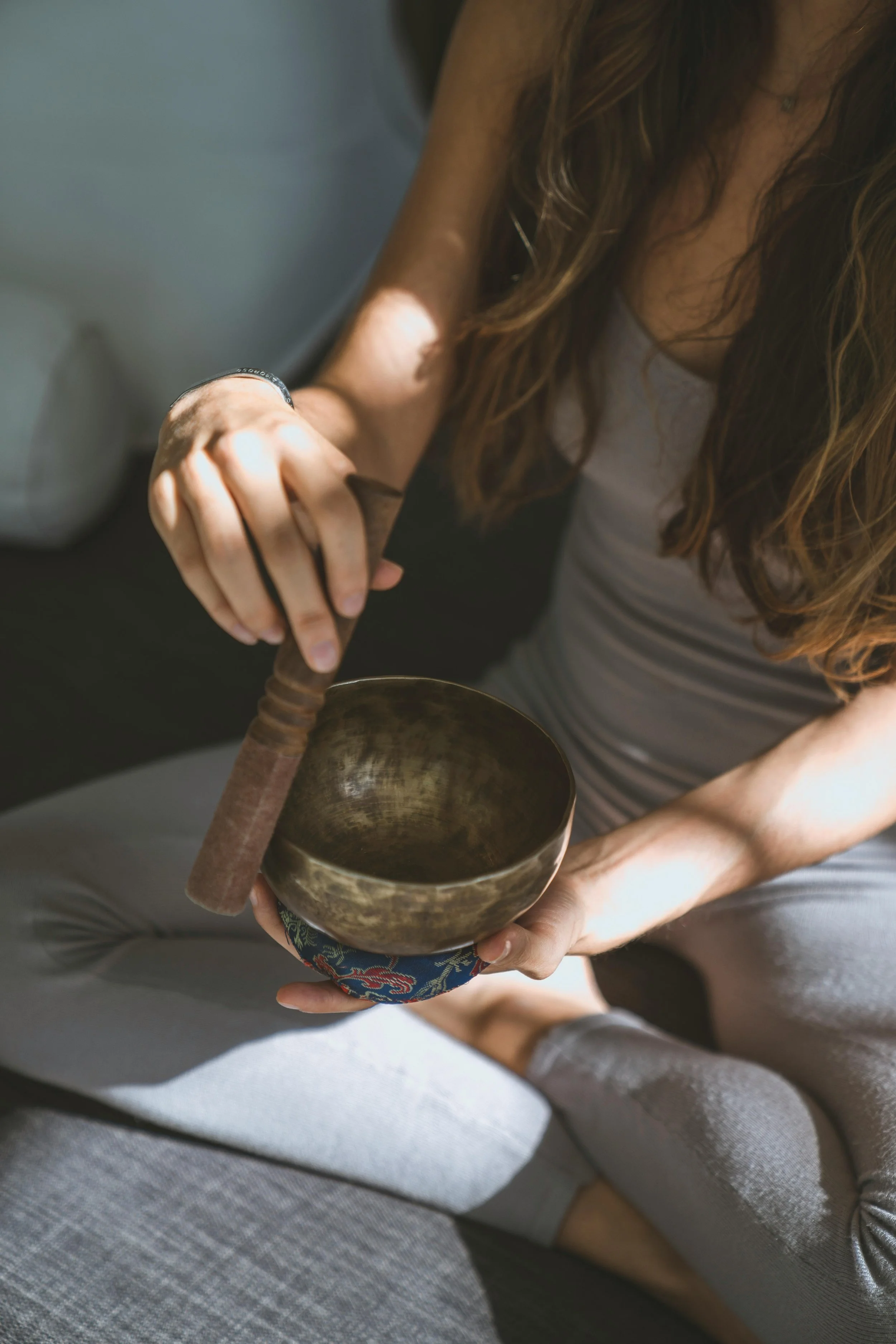 A woman with long brown hair sitting cross-legged, holding a Tibetan singing bowl in her left hand and a wooden mallet in her right hand, using it to play the bowl.