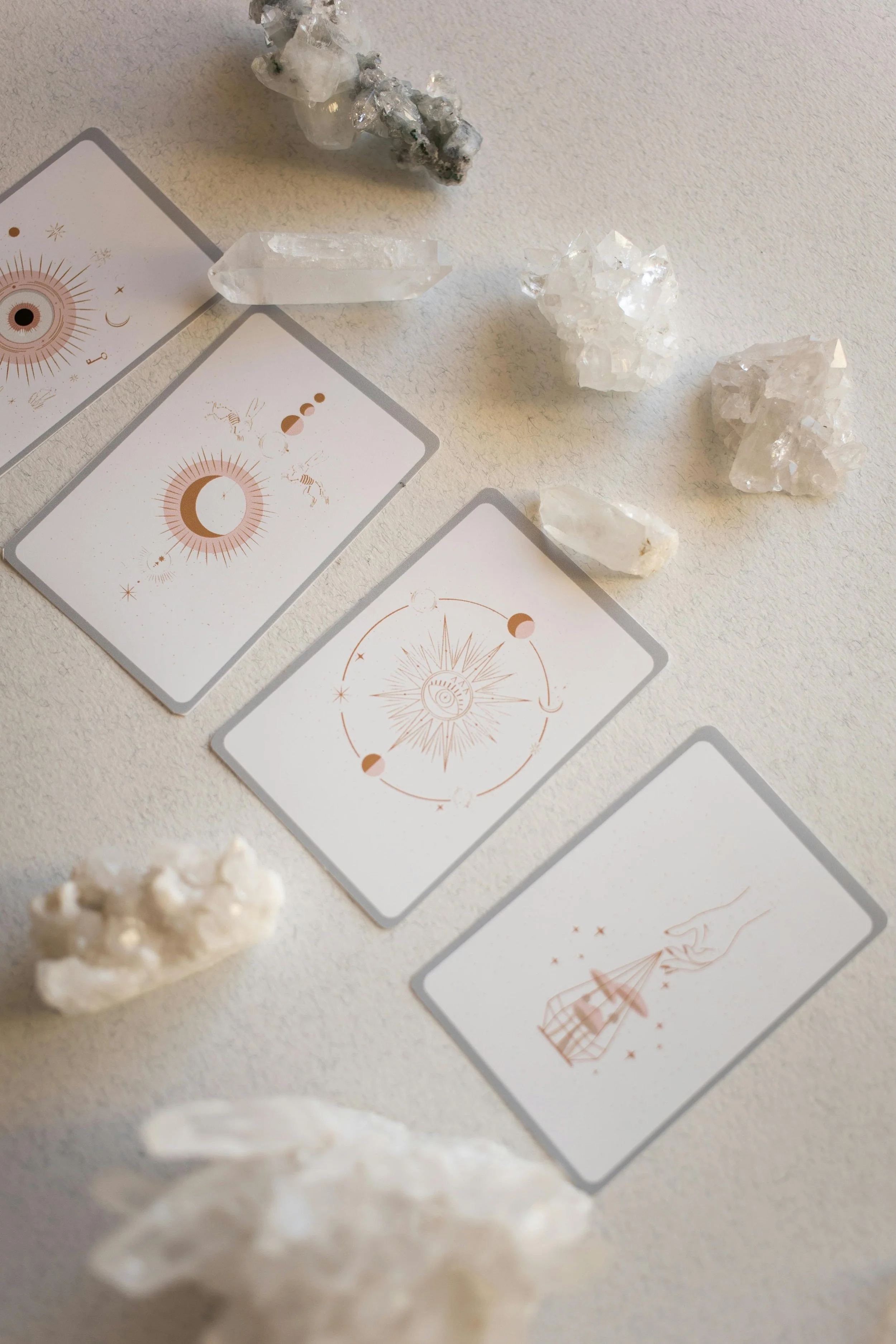 Four tarot-like cards with mystical symbols, surrounded by white crystals and mineral stones on a plain white surface.