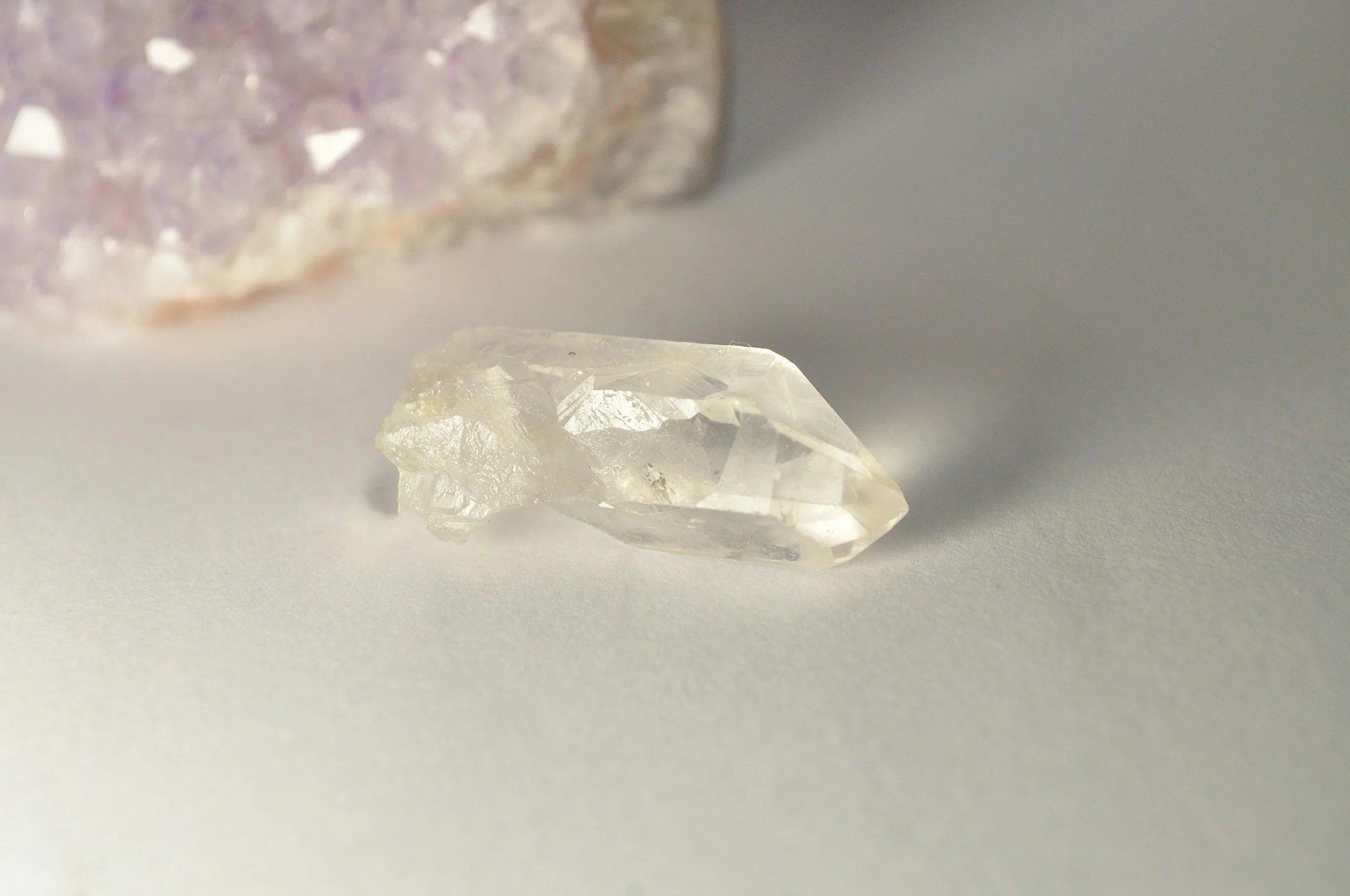 A raw, uncut clear quartz crystal with a shiny, faceted surface, lying on a plain, light-colored surface with a blurry, textured stone in the background.