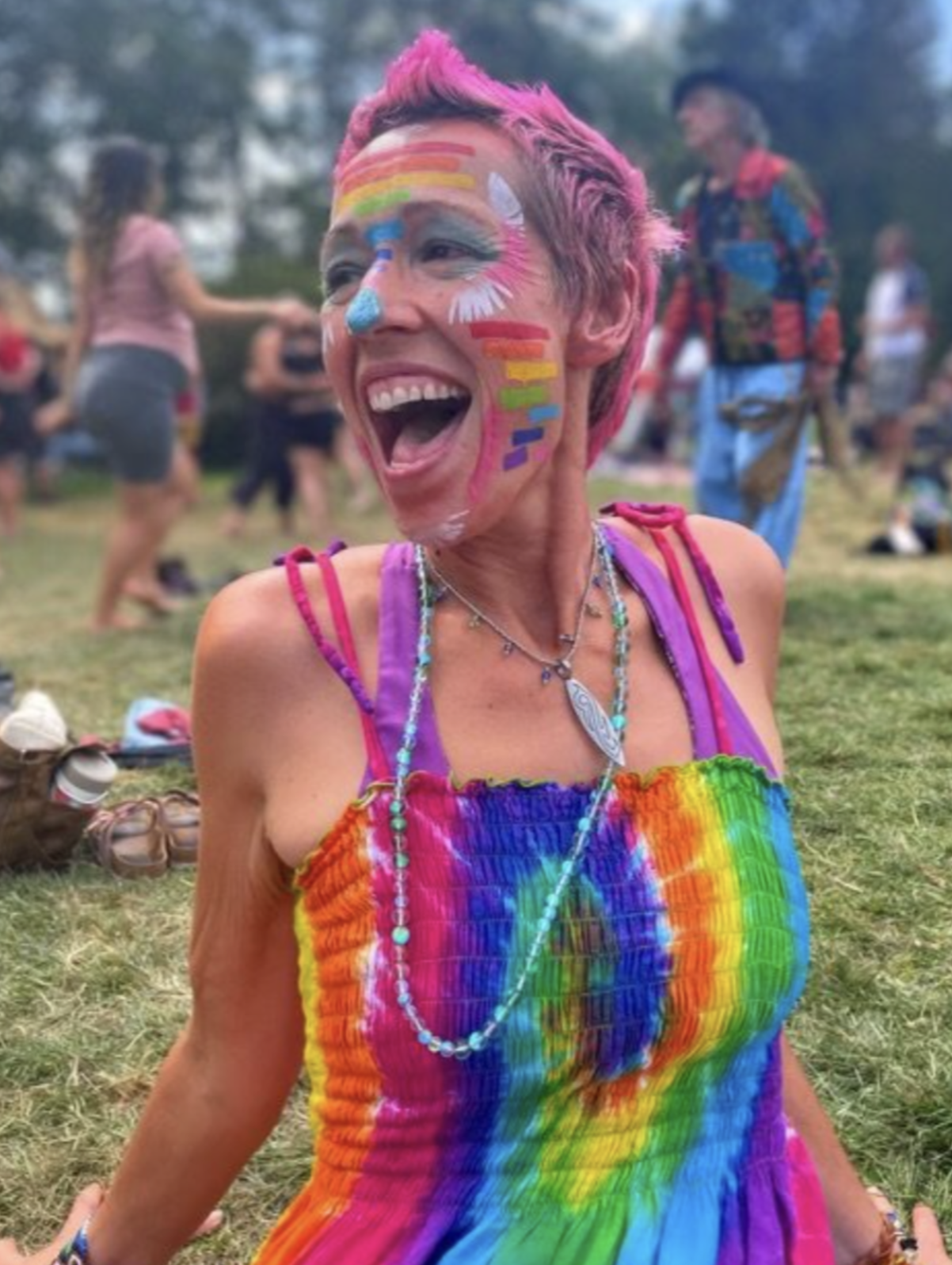 Woman with face painted with rainbow colors, wearing a rainbow tie-dye dress, at an outdoor festival or gathering, smiling and laughing.