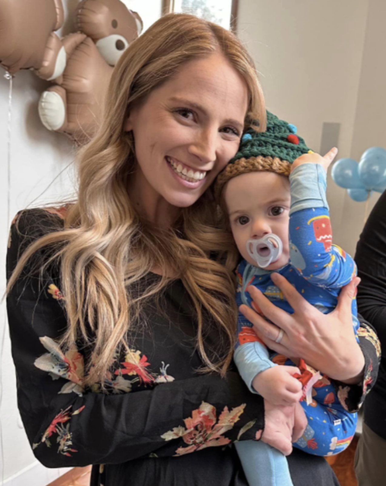A smiling woman with long blonde hair holding a baby wearing a colorful elf hat and blue pajamas with festive patterns, in a decorated room with balloons and teddy bear-shaped balloons in the background.