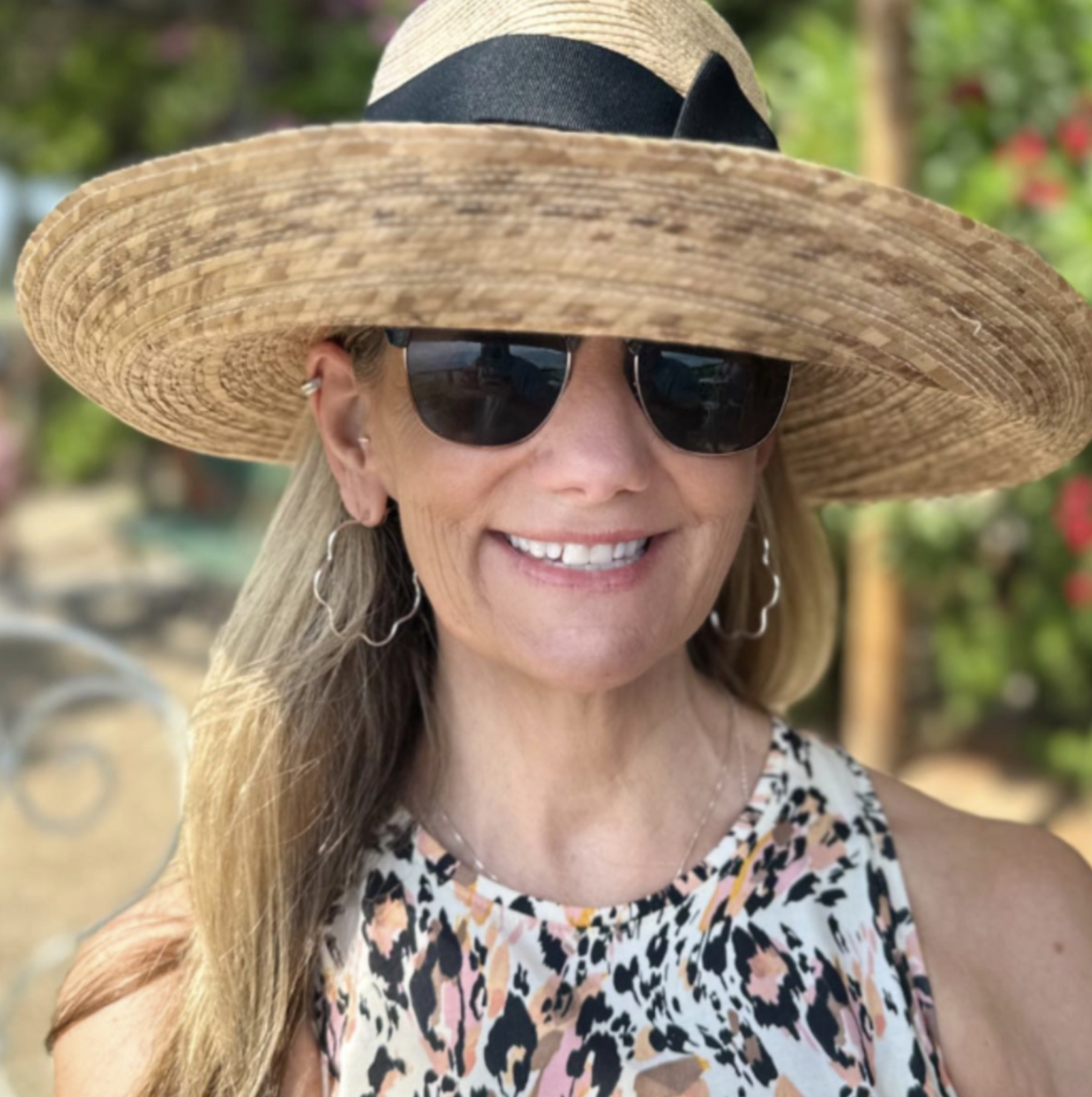 Woman smiling outdoors, wearing a wide-brimmed straw hat, black sunglasses, and a sleeveless animal print top, with a blurred background of greenery and flowers.