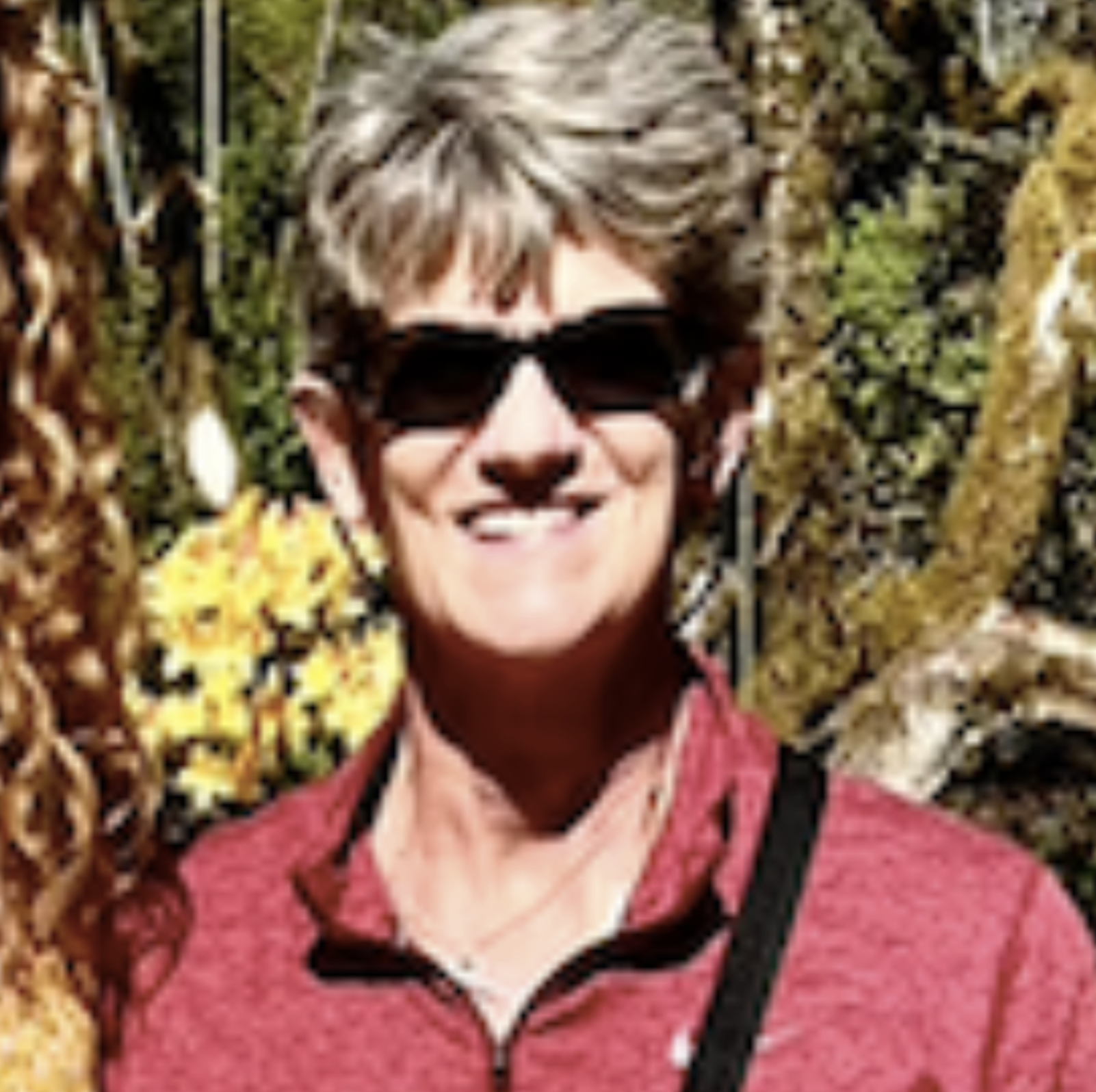 A smiling woman with short gray hair wearing sunglasses and a pink jacket standing outdoors with greenery in the background.