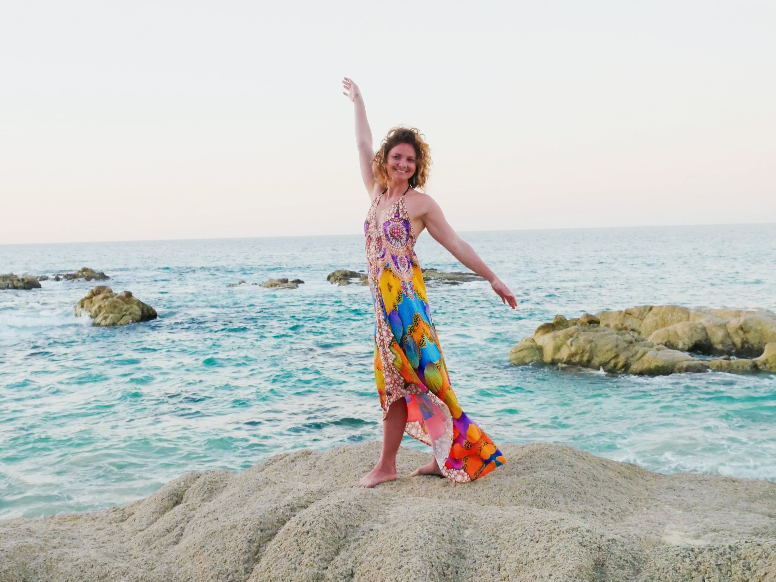 Woman in a colorful, patterned dress dancing and smiling on rocks at the beach with the ocean in the background.