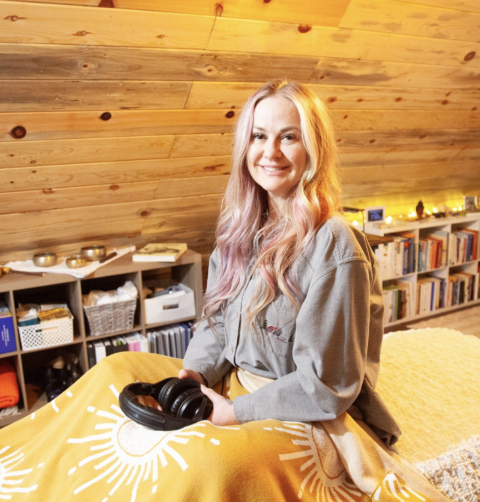 A woman with long, wavy, blonde hair smiling while holding a pair of black headphones in a cozy, artfully decorated room with wooden walls, a skeleton model, bookshelves, and a bed with a yellow blanket.