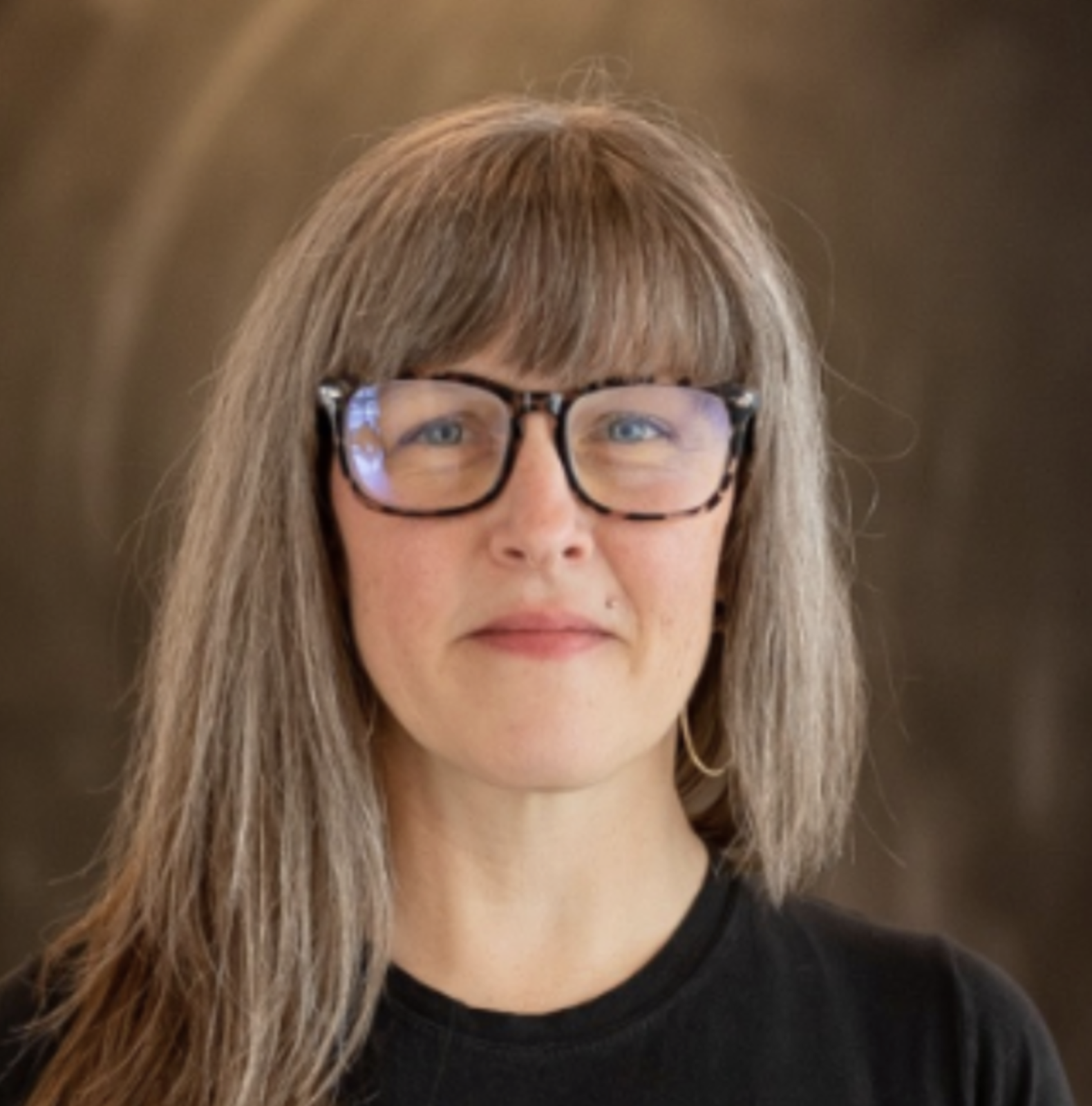 A woman with long gray hair, glasses, and a black shirt standing indoors against a dark background.