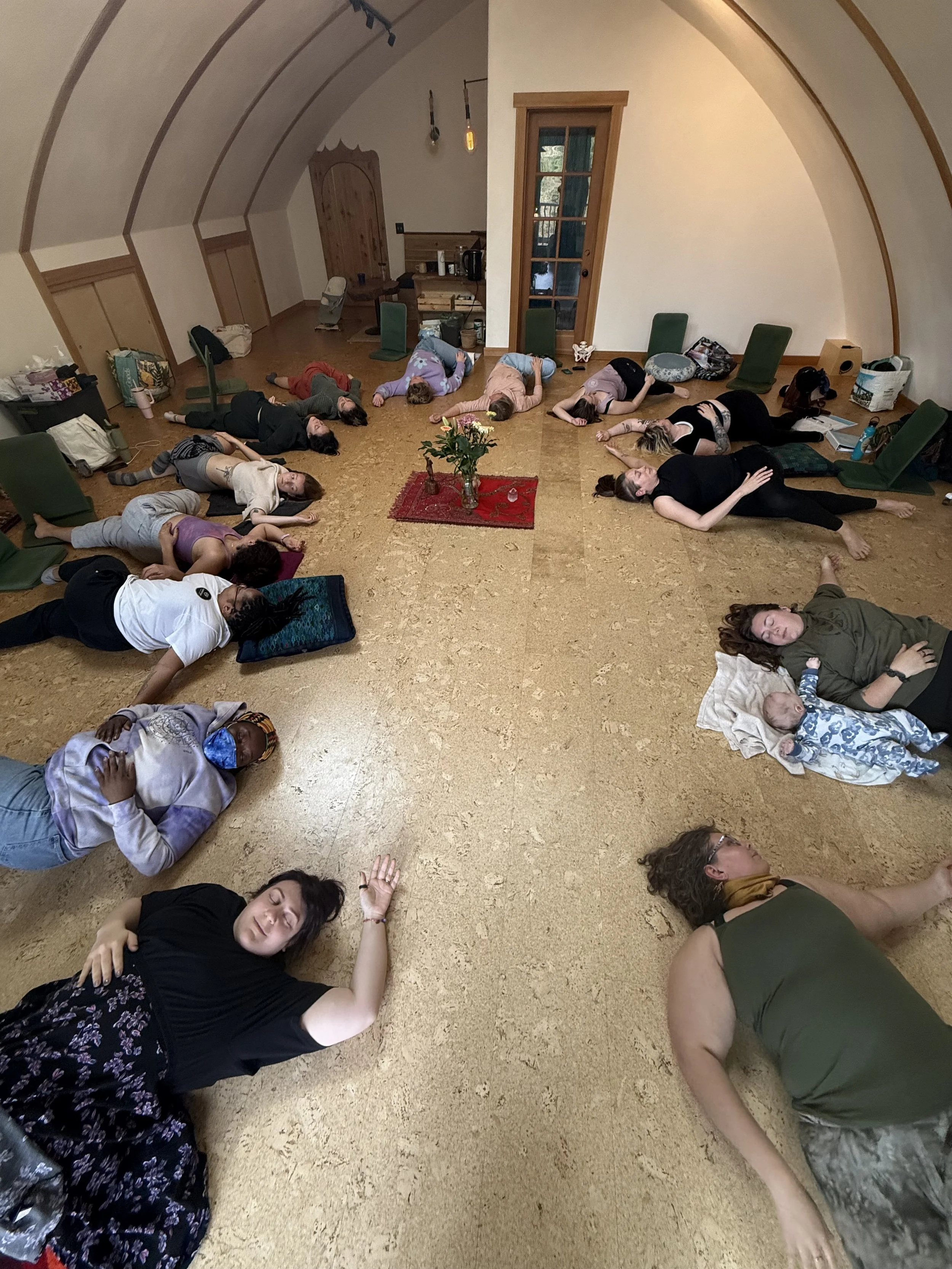 Group of people lying on the floor in a circle during a workshop or meditation session in a cozy room with a rounded ceiling.