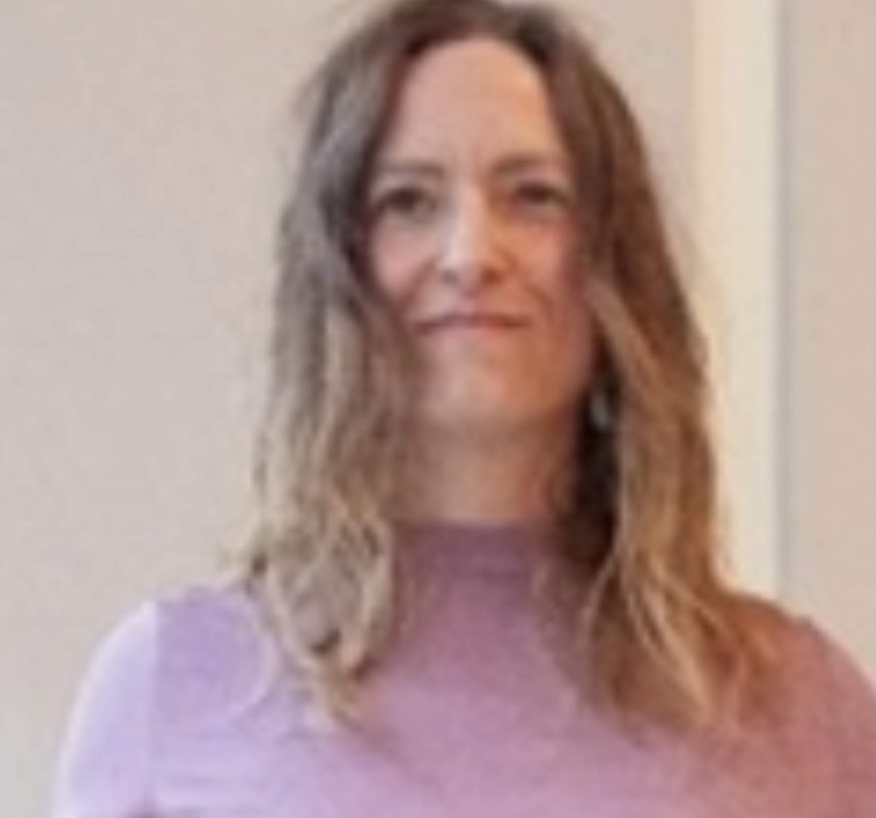 A woman with shoulder-length wavy hair, wearing a light purple top, standing against a plain wall.