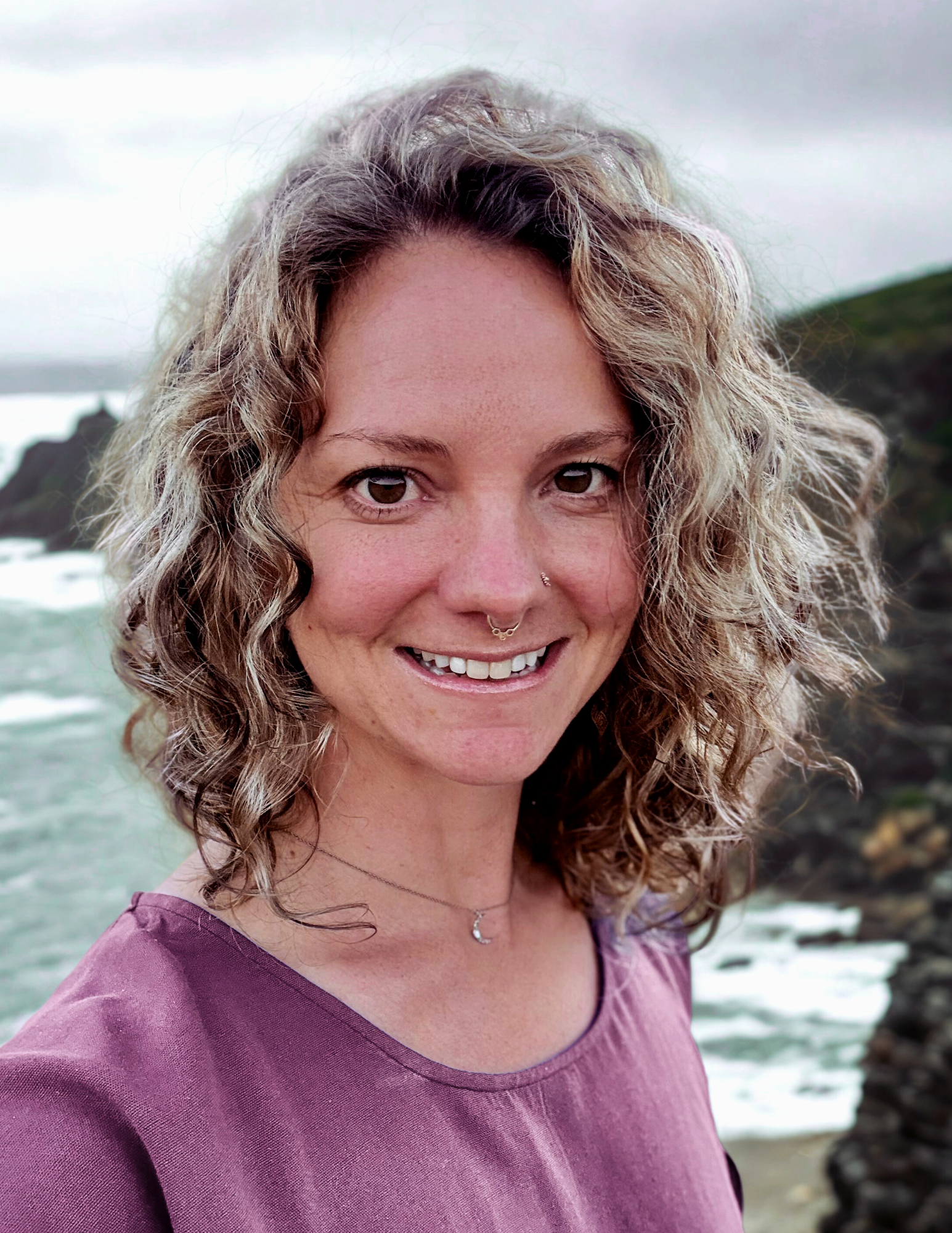 Smiling woman with curly hair, wearing a purple top, standing near a rocky shoreline with the ocean and cloudy sky in the background.