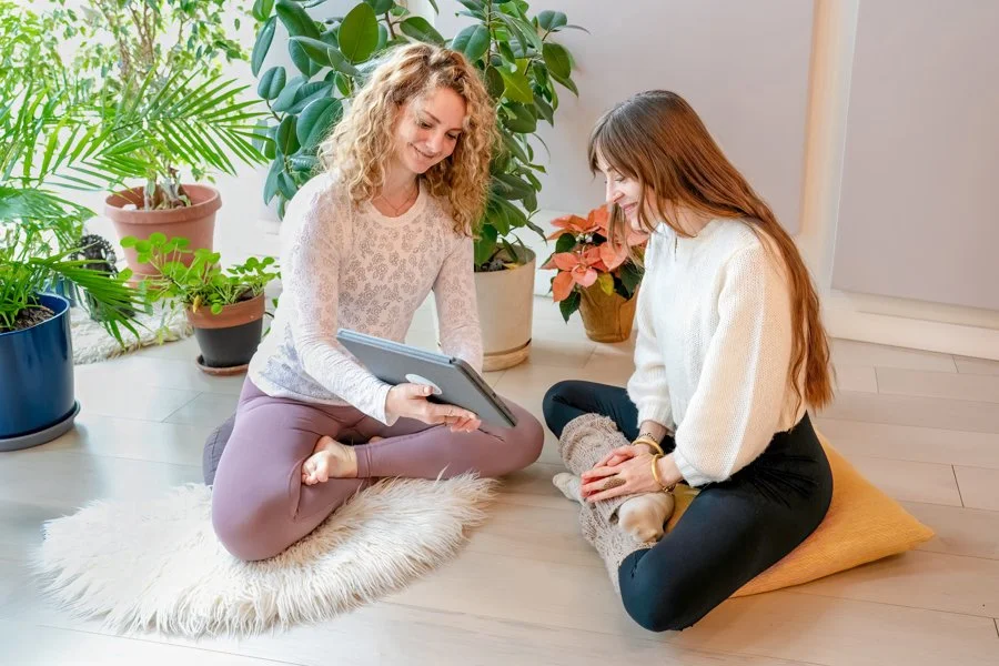Two women sitting on the floor near houseplants. One woman is holding a tablet, and both are smiling. One woman is sitting on a fluffy white rug, and the other is on a cushion. The setting appears to be a cozy indoor space.