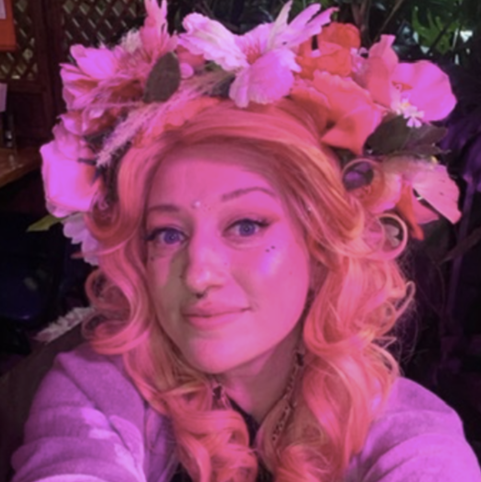 Woman with long, curly blonde hair and blue eyes wearing a floral crown of large pink and peach flowers, in a setting with pink lighting.