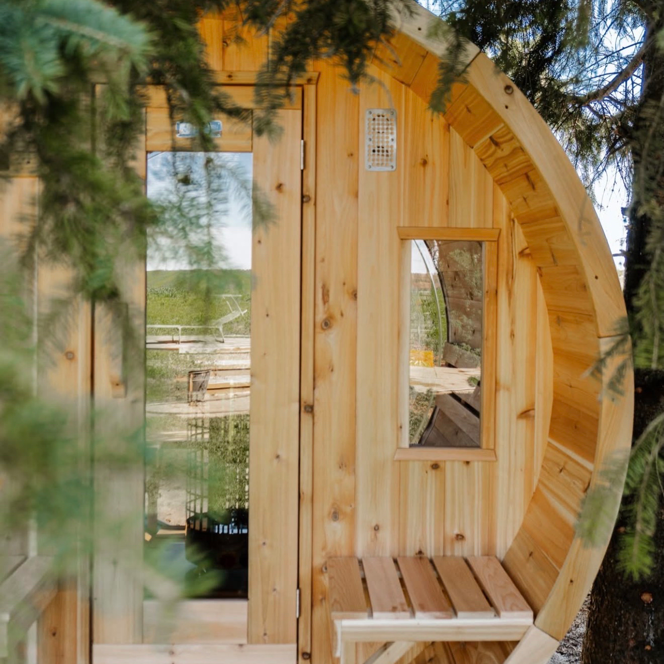 A small wooden yurt with a door and a window, surrounded by trees and greenery