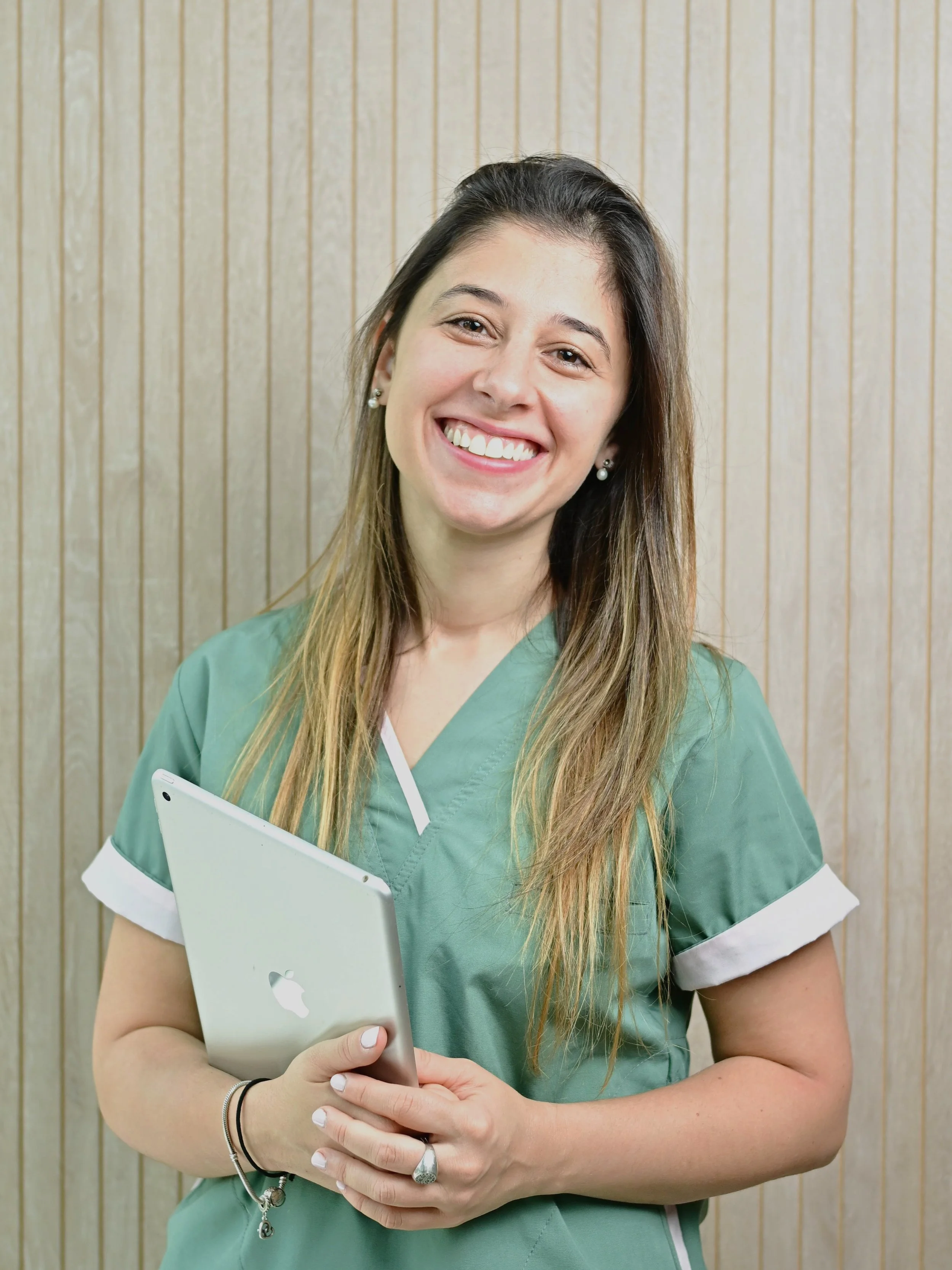 Enfermera sonriente con uniforme verde y sosteniendo una tableta plateada, de pie frente a un fondo de madera panelada.