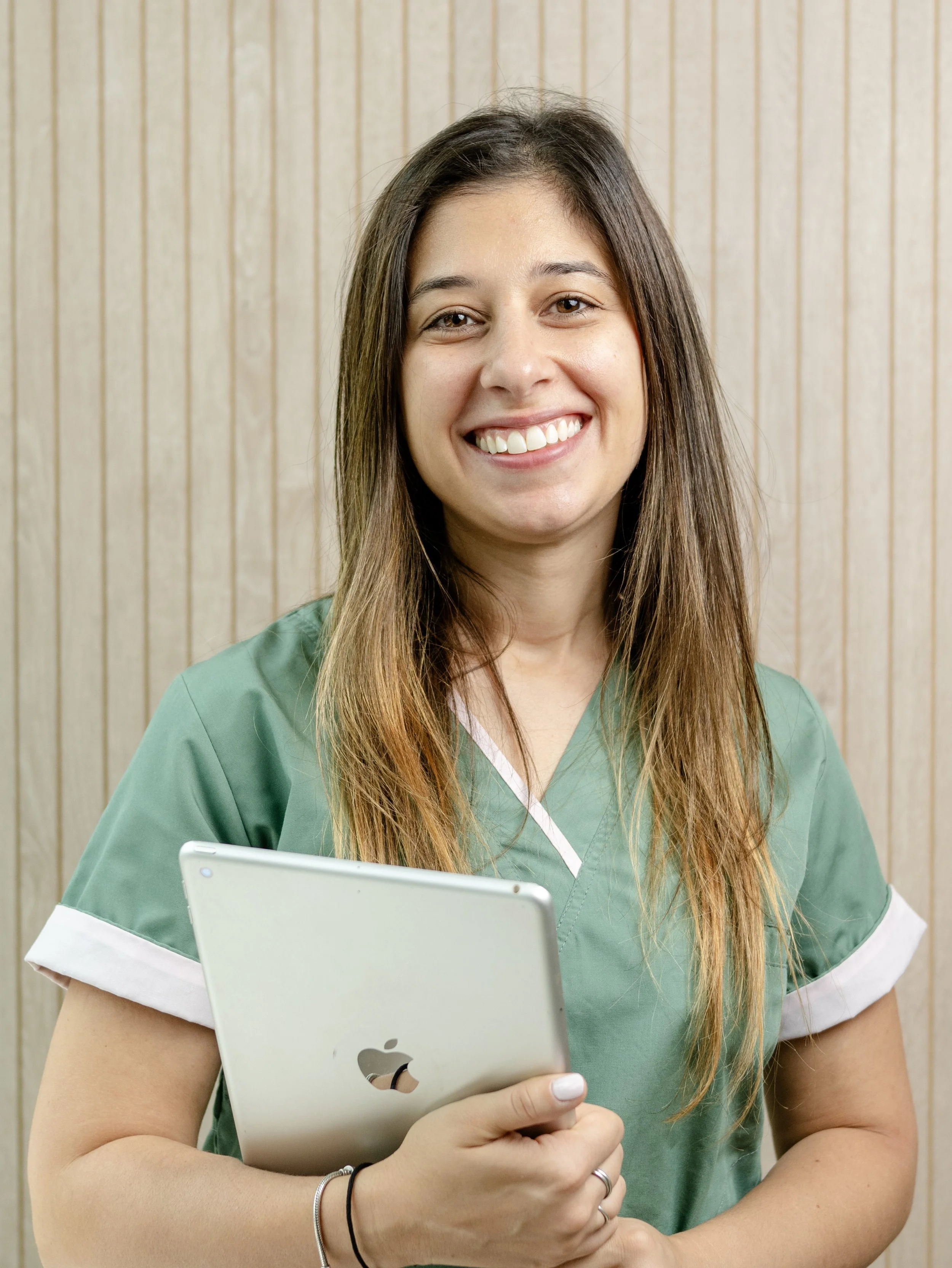 Mujer con uniforme médico sosteniendo una tableta, sonriendo y frente a fondo de madera.