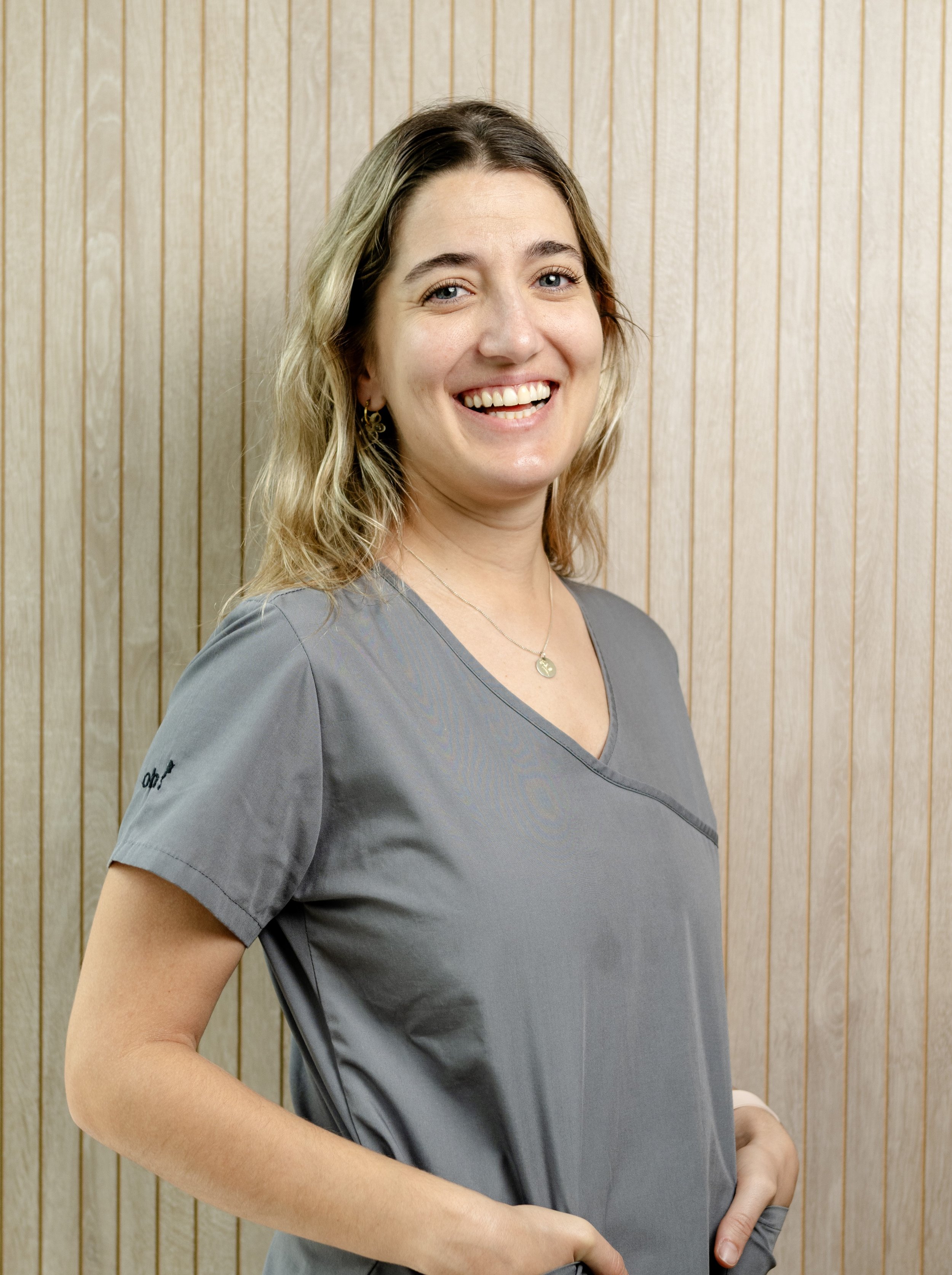 Mujer sonriendo con uniforme gris, de cabello rubio y ondulado, de fondo con paredes de madera.