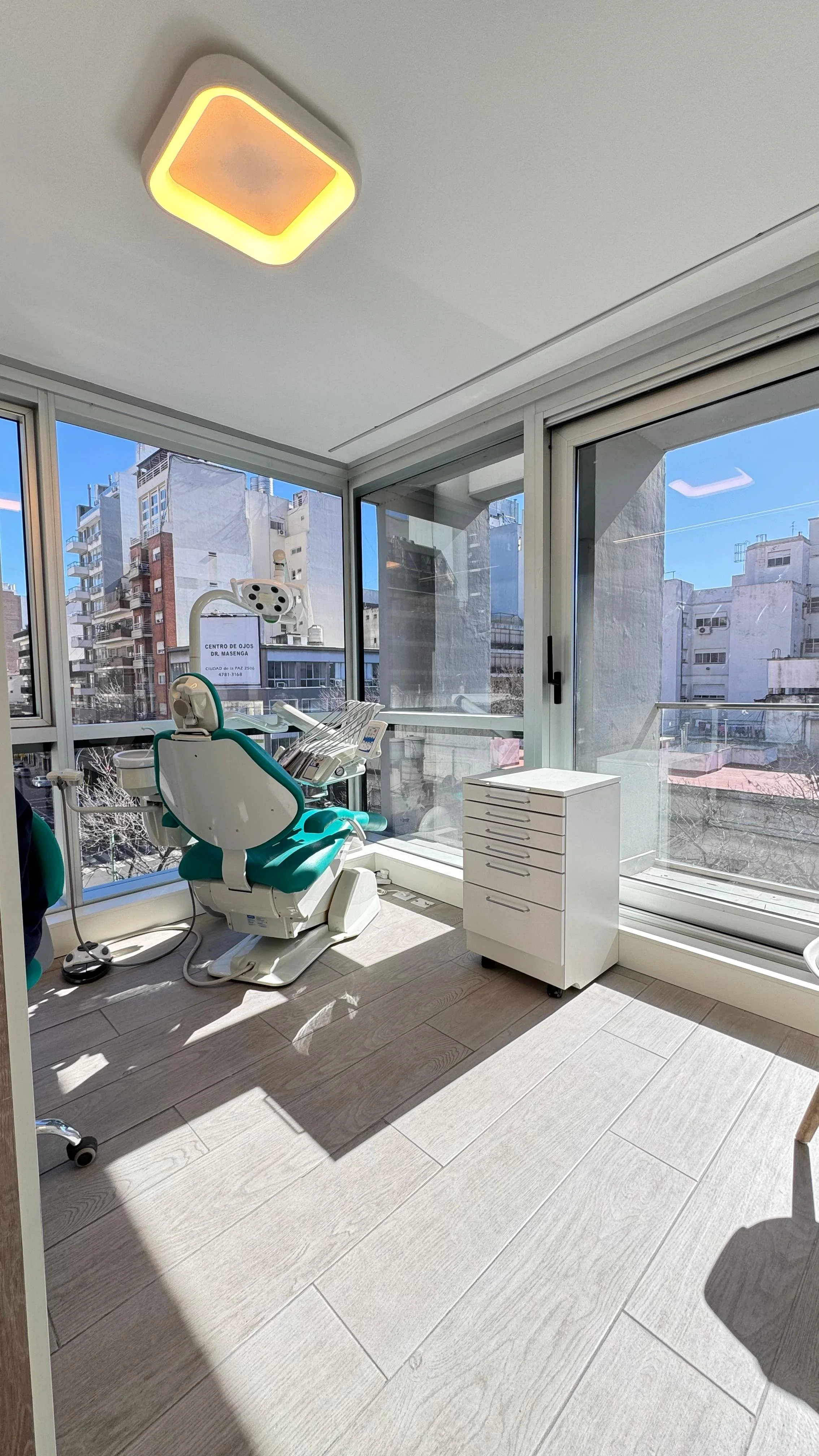 Consultorio odontológico con sillón dental, equipo dental, ventanas grandes con vista a edificios, iluminación moderna en el techo y suelo de madera clara.