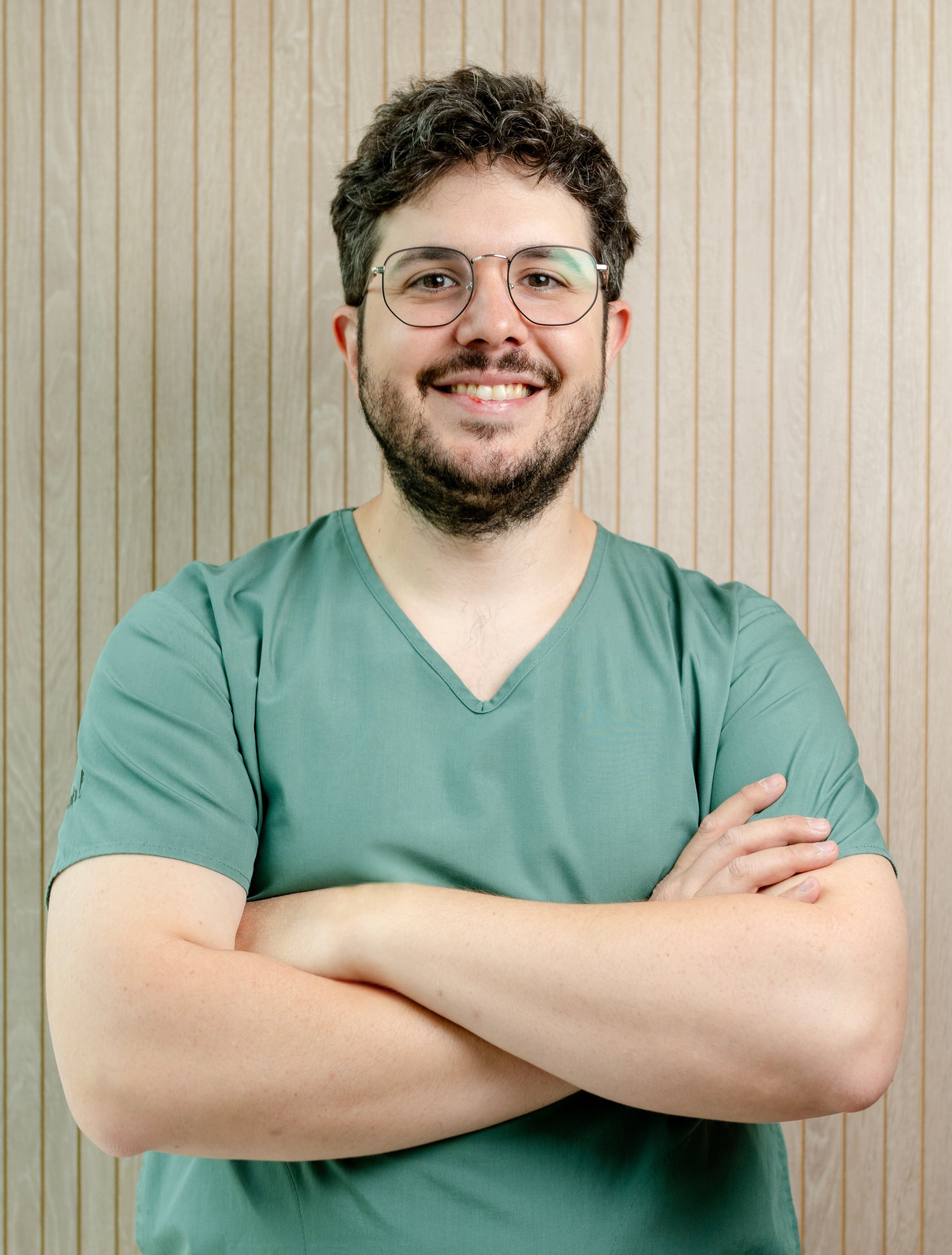 Joven hombre con barba y gafas, sonriendo, con brazos cruzados, en una camisa verde, frente a una pared de madera.
